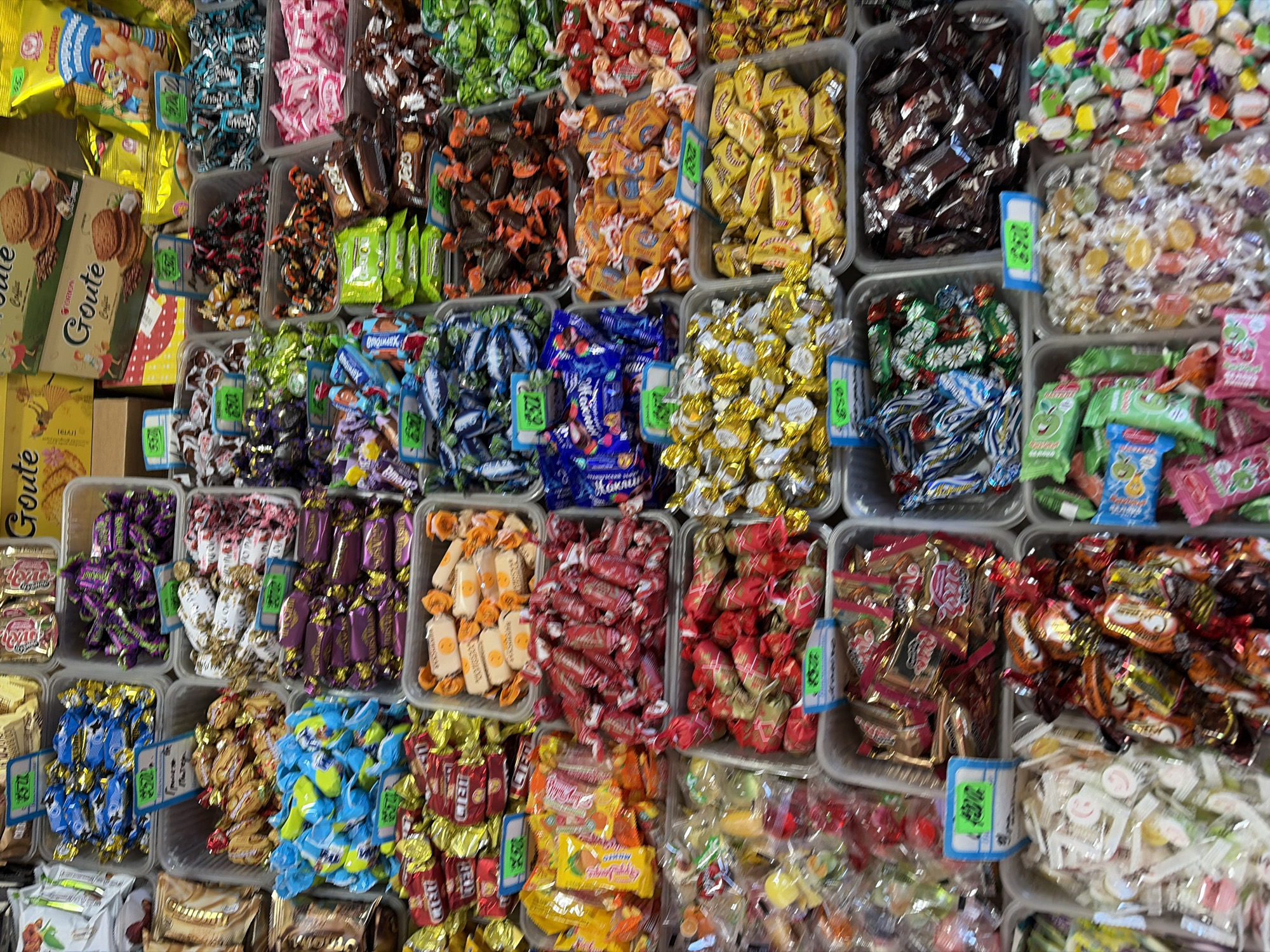 This media file captures a vibrant, high-angle view of a diverse candy and cookie display, likely in a market or shop in Sochi, Russia. The scene is dominated by numerous plastic bins filled with a vast assortment of individually wrapped candies, sweets, and some packaged cookies.
The display is arranged in a grid-like pattern, with various confectionery items grouped by type and color. Candies come in an array of wrappers, including shiny metallics, bright reds, blues, greens, yellows, purples, pinks, and browns, creating a rich tapestry of colors and textures. Many bins have small, blue-rimmed price tags with green inserts, indicating prices in Cyrillic script.
Notable visible items include:
* **Cookies/Biscuits:** On the left, several boxes and bags of "Goute" brand cookies are visible, some labeled "Caramelise" and "Original".
* **Candies:** An extensive variety of wrapped candies fills the bins. Some wrappers appear to be for caramel, nougat, hard candies, and chocolates. Specific Cyrillic text observed on wrappers includes "Оригинал" (Original) on a blue wrapper, "Красный Октябрь" (Krasny Oktyabr - Red October) on a red wrapper, "Алёнка" (Alyonka) on a brown wrapper, "Апельсиновый" (Apelsinovy - Orange) on a yellow bag, and "Вишня" (Vishnya - Cherry) on a pink bag. Other identifiable shapes resemble popular international brands like Kit Kat.
* **Pricing:** Price tags clearly show numbers such as "250", "220", "200", "280", "150", "230", "170", and "240", presumably indicating costs per 100 grams or per item in Rubles.
No people are visible in the image, and the time of day or weather cannot be determined. The focus is entirely on the colorful and abundant display of confections ready for sale.