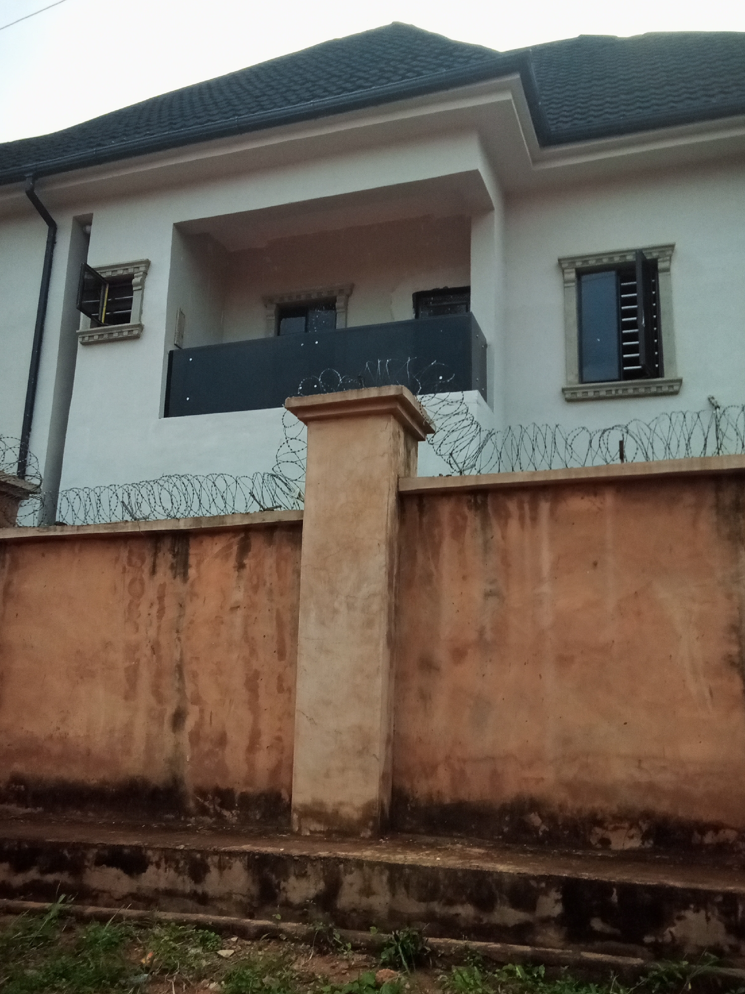 This image shows a residential building located in Oko Town, Nigeria. The building has a light-colored exterior and a dark-tiled roof. There are windows with dark frames and louvers, some of which are open. A balcony with a dark railing is visible on the upper floor. In front of the building, there is a concrete wall topped with barbed wire. A pillar with a textured surface stands in front of the wall. The area in front of the wall has some green vegetation. The overall scene depicts a secured residential property.