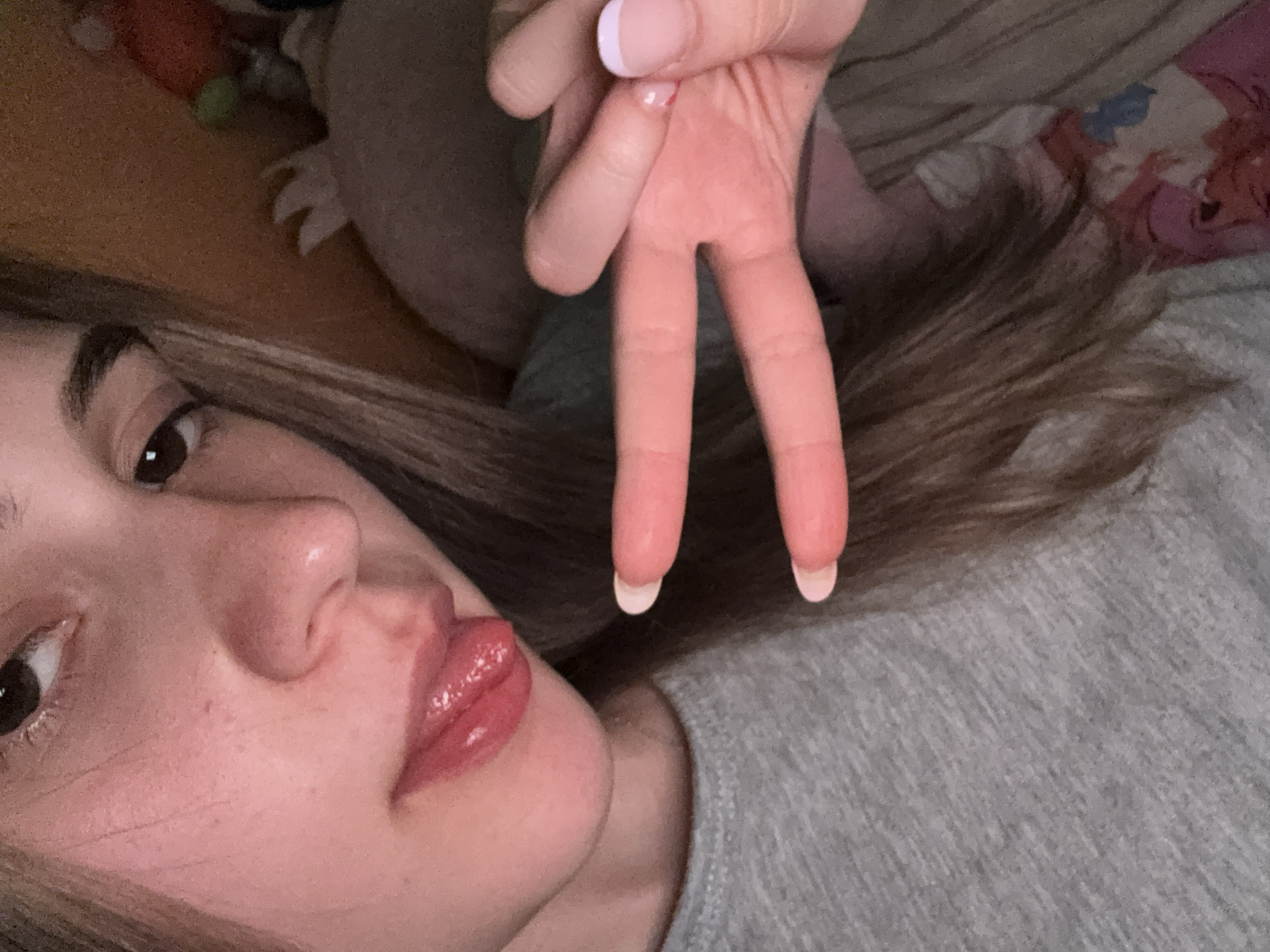 A close-up, slightly angled, indoor selfie of a young woman with light brown hair. The woman is lying on what appears to be a grey t-shirt or bedding. Her face is prominent, showing her eye, nose, and glossy lips. She is holding up her hand in a peace sign near her face. The fingertips of the peace sign have white nail polish. The background is blurred, showing what might be a bed with some stuffed animals. Location information is unavailable.