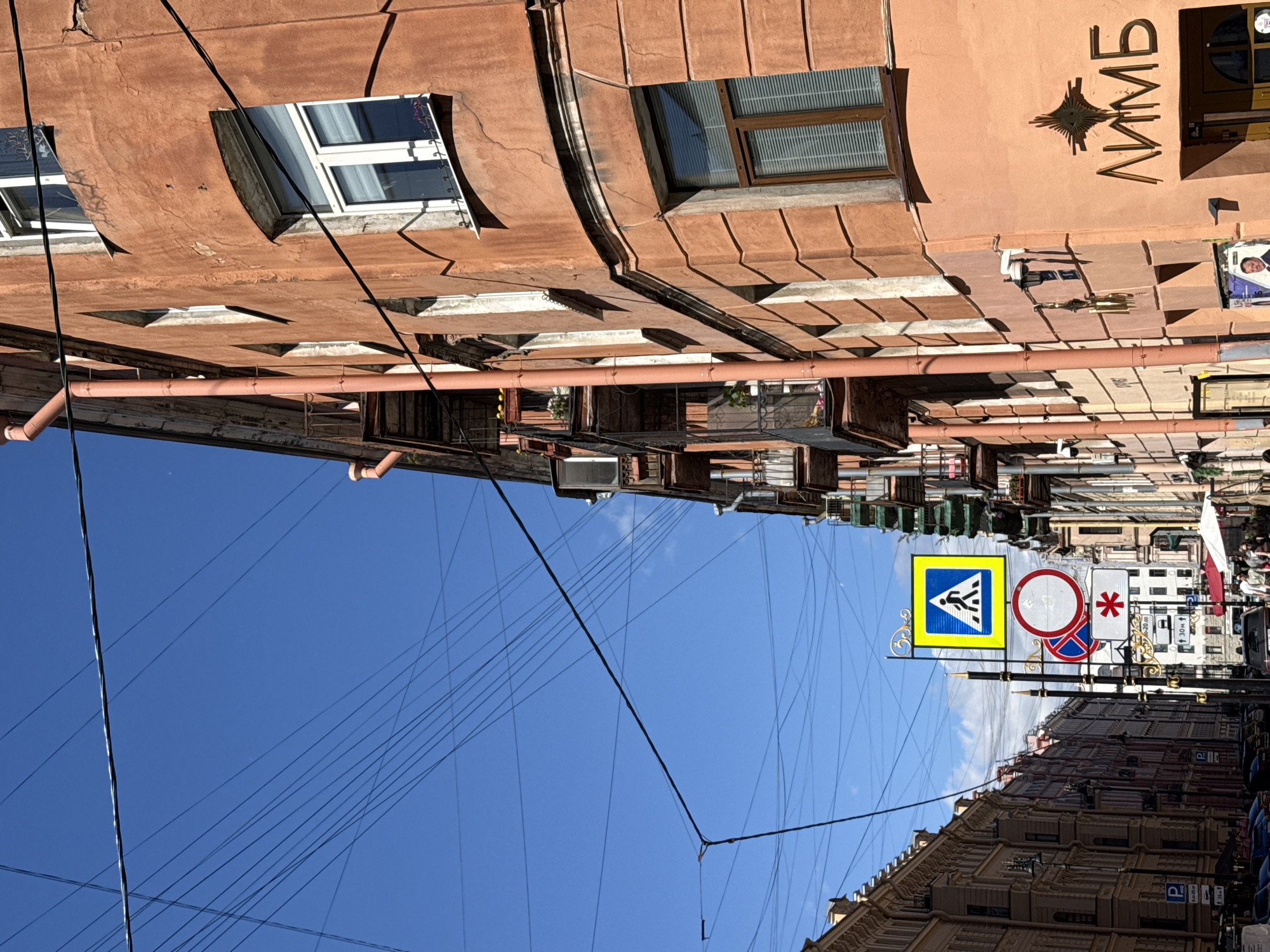The image depicts a street scene in Saint Petersburg, Russia, viewed from a low angle looking upwards. The perspective is skewed, with the buildings and sky appearing rotated. The buildings are an ochre color, with visible cracks and peeling paint on their facades. Several windows are visible, some with white blinds and others with wooden louvers. A large, pinkish-red pipe runs horizontally across the middle of the image, with smaller pipes branching off it.
In the sky, a complex network of black electrical wires crisscrosses the frame, creating a dense web against the bright blue sky. In the lower right portion of the image, a pedestrian crossing sign is visible, a yellow square with a blue border and a white silhouette of a person walking. Next to it are two circular traffic signs, one with a red border and a diagonal red bar, and another with a red asterisk. Further down the street, more buildings are visible, some appearing ornate with detailed architectural features. The time of day appears to be sunny, with shadows cast by the buildings and signs. There is no discernible activity of people actively engaged in anything, but the overall impression is of a lived-in urban environment. The word "ЛИМБ" is visible in large, stylized metallic letters on one of the building's upper walls.