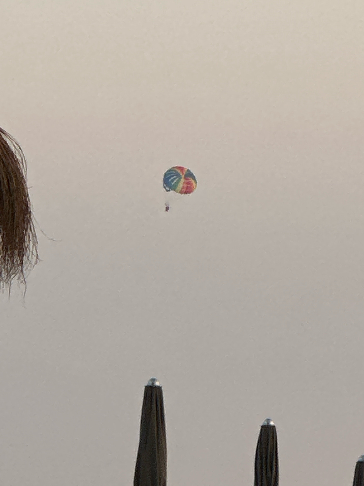 The image is a close-up shot of a paraglider against a pale sky. The paraglider, which is colorful with red, blue, yellow, and green segments, is small in the frame and appears to be high in the sky. A person is attached to the paraglider, though very small and difficult to discern details. In the foreground, the top portion of three dark, umbrella-like objects are visible, suggesting they are positioned outdoors, possibly at a beach or resort. The umbrella tops are pointed and have a metallic silver cap. To the left edge of the frame, a section of dark, long hair is visible, obscuring a portion of the background. The overall scene suggests an outdoor recreational activity taking place in a coastal environment, possibly in Rafailovići, Montenegro, given the presence of beach umbrellas and the activity of paragliding often associated with tourist areas.