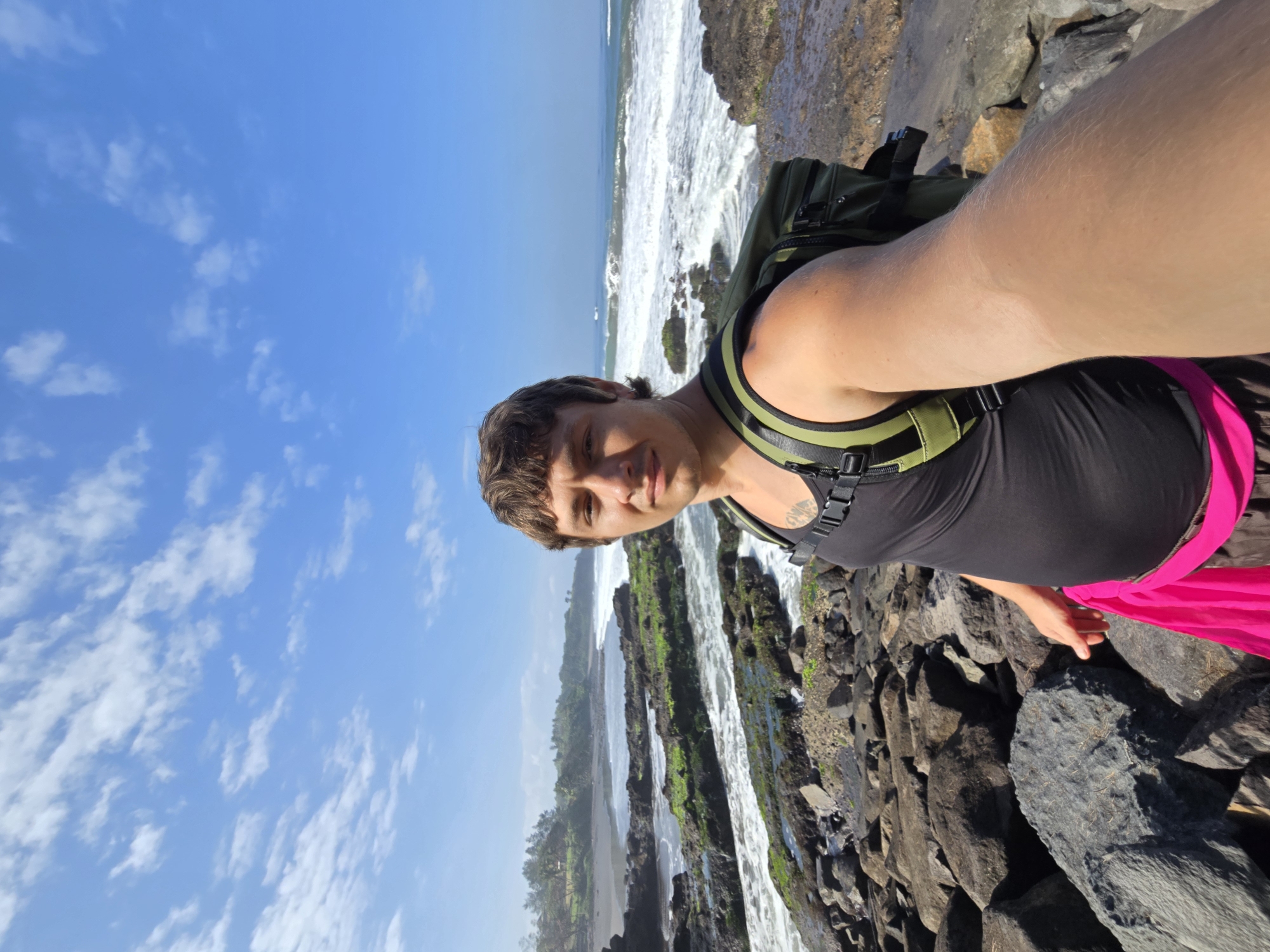 A man is taking a selfie on a rocky coast with waves crashing. He is wearing a black tank top, a green backpack, and pink shorts. The background shows a black sand beach, lush green vegetation, and a clear blue sky with scattered clouds. The man appears to be in his late 20s or early 30s, with short brown hair and a light complexion. The rocky terrain is covered in dark stones and patches of green algae. The ocean water is a vibrant blue with white foam from the waves. This scene is likely located in the city of Selemadeg, Indonesia, known for its scenic coastal areas.