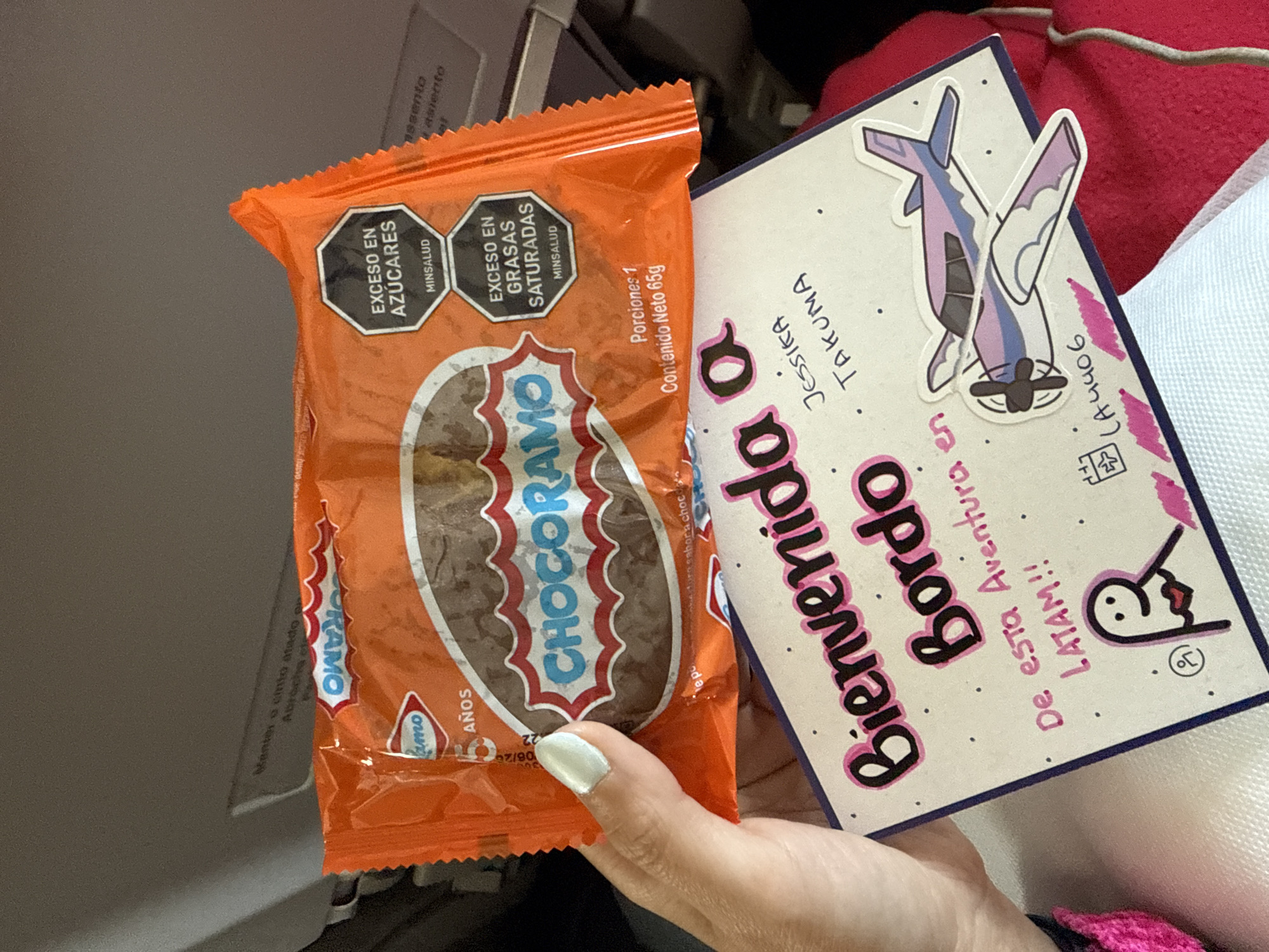 A person is holding a Chocoramo snack and a welcome card. The snack is in an orange wrapper with the words "CHOCORAMO" in a red and white logo. The wrapper also has warning labels regarding excess sugar and saturated fats, in Spanish. The welcome card is light pink and decorated with dots. It has the words "Bienvenida a Bordo" (Welcome Aboard) in large letters, along with the names "Jessica" and "Takuma", and the flight number "LA4406". The card also has a sticker of a cartoon airplane. The person's hand with a light blue nail polish is visible. The background appears to be the interior of an airplane. The image does not depict any location in the city of Orlando, Florida, United States of America.