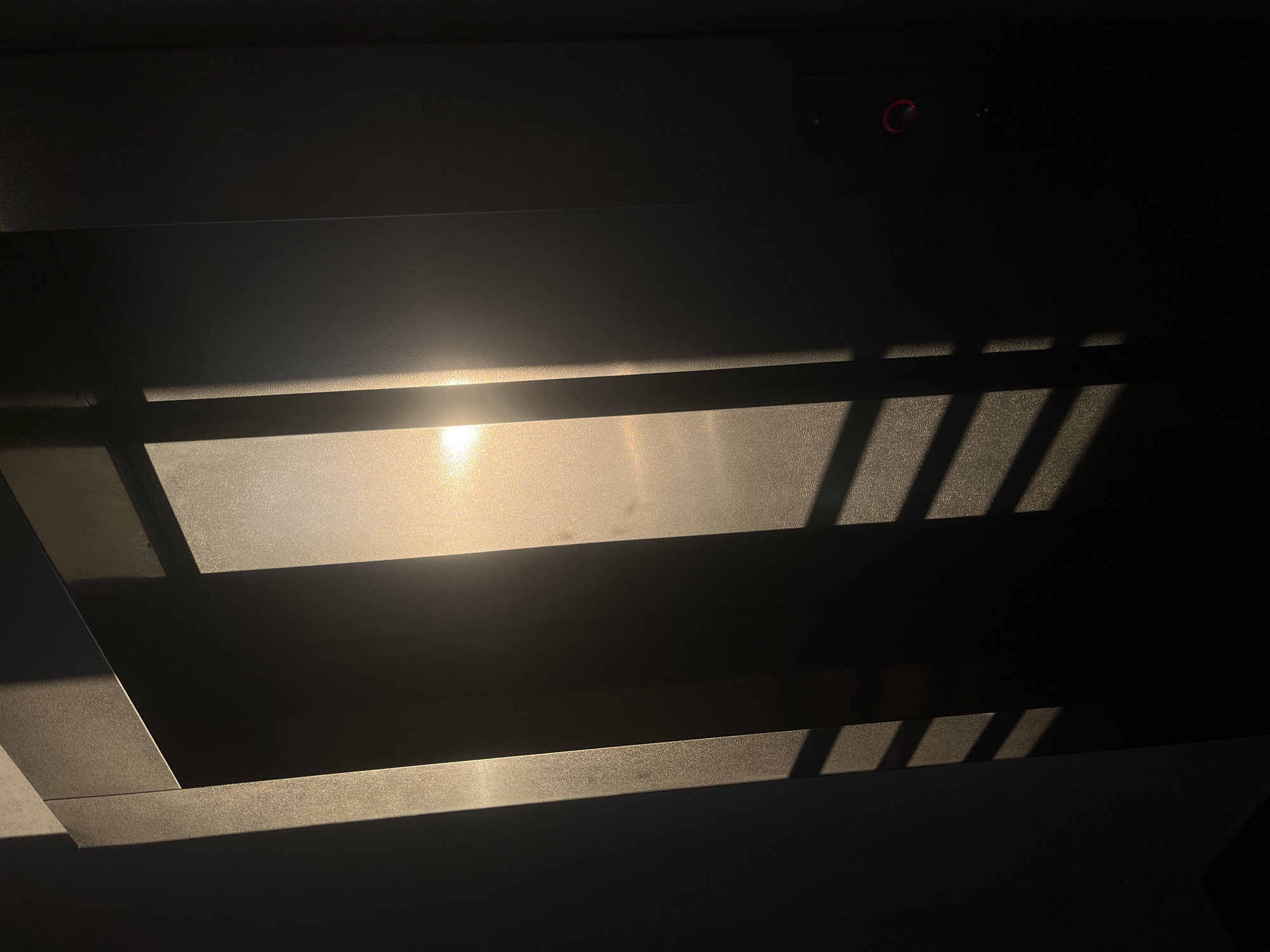 The image shows a shadowed interior space with sunlight streaming through an opening, casting linear patterns of light and shadow on the walls and ceiling. The light source is not directly visible but creates a bright, diffuse glow where it hits the surface. Above the area of light are two small dots that look to be electronic and a small red circle. The angle and stark contrast between light and dark suggest a strong, direct source of light, possibly a window. There is no evidence to suggest the location of Saint Petersburg, Russia.