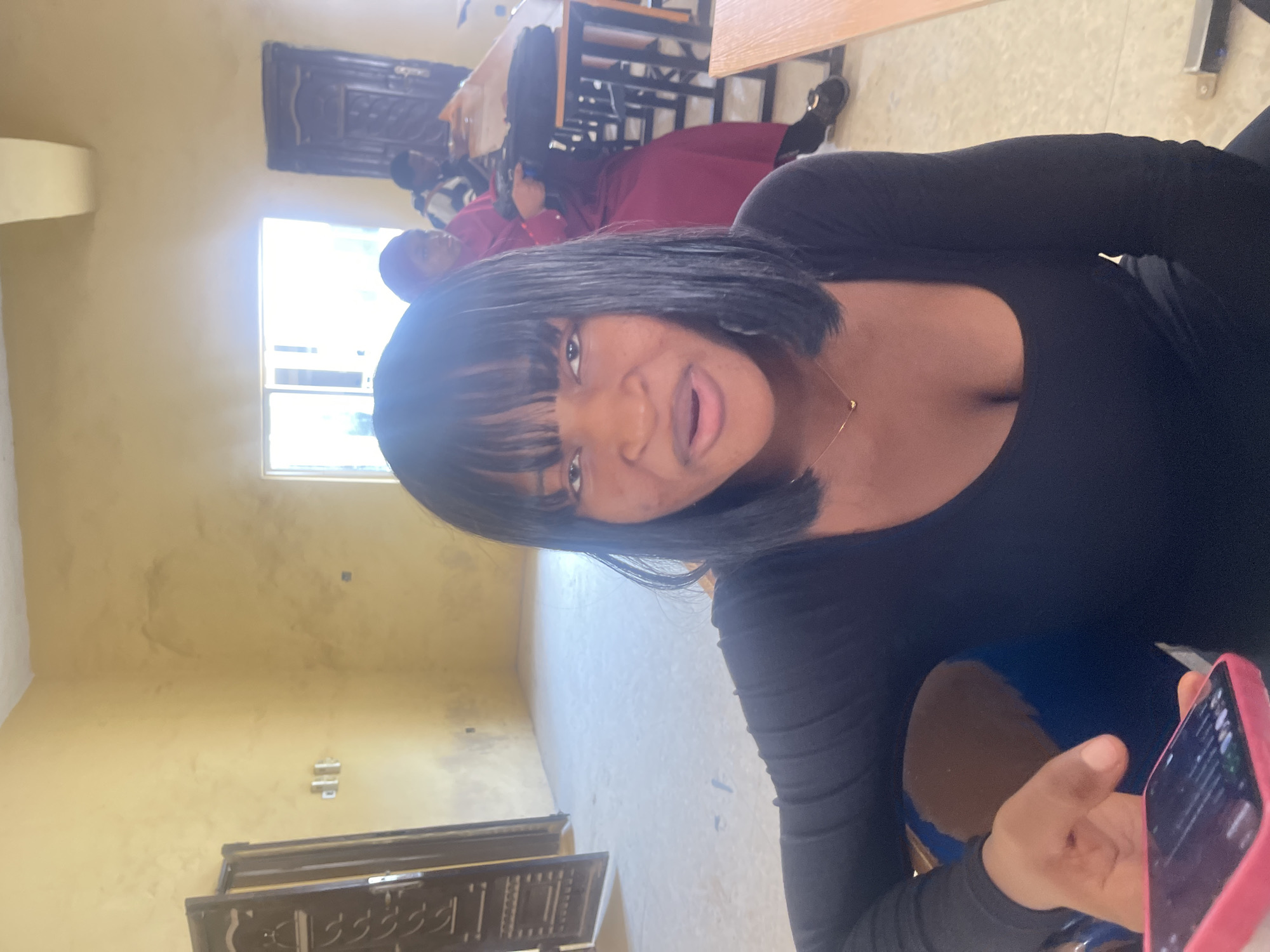 A close-up, waist-up shot shows a young Black woman with straight black hair and bangs, wearing a black long-sleeved shirt, and a thin gold necklace with a heart pendant. She is looking directly at the camera with her mouth slightly open. Her left hand is visible in the foreground, holding a pink cell phone. In the background, out of focus, are two other people, one wearing a red hijab and maroon dress, and another person wearing a striped sweater. The scene appears to be an indoor classroom or meeting room with beige walls and a concrete floor. Sunlight streams through a large window on the far wall. The environment is consistent with a building in Rafin Par, Nigeria, possibly a school or community center.