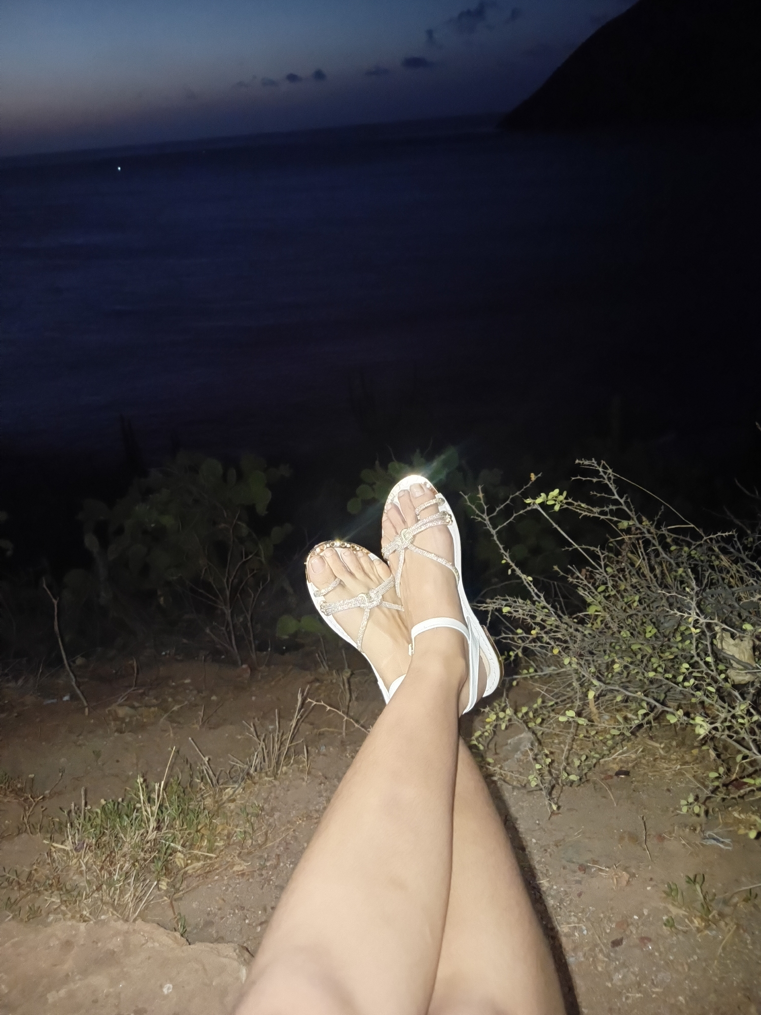 A person's legs are crossed, clad in gold and silver flat sandals with a white sole, resting on an elevated position overlooking a dark sea. The background includes a night sky and a faint light on the horizon. The location is likely Juan Griego, Venezuela, given the coastal landscape. Vegetation, including small shrubs and grass, is visible along the edge where the person is situated. The ground appears to be dirt or gravel. The overall scene is dim, possibly at dusk or night.