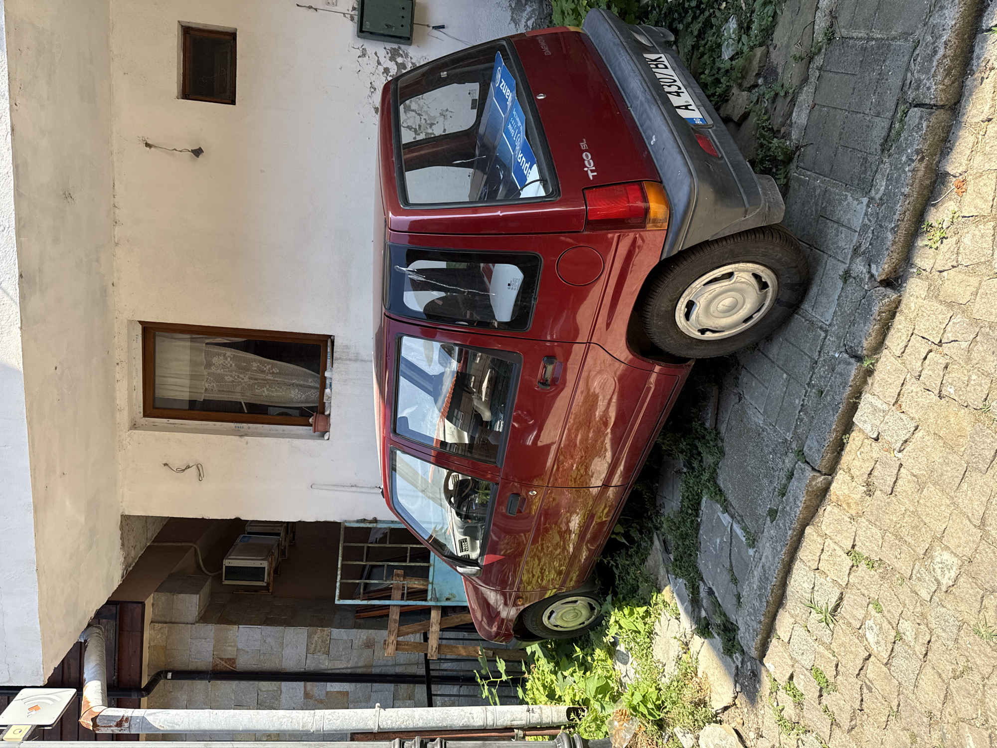 A red Daewoo Tico SL is parked on a cobblestone street in Sozopol, Bulgaria. The car is positioned at an angle, with its rear facing the viewer and slightly to the right. The vehicle is surrounded by overgrown greenery and appears to be in a somewhat neglected state. A blue sticker with text is visible on the rear window, as well as a license plate that reads "A 4307 BK". Behind the car, there is a white building with a window and a small green electrical box mounted on the wall. To the left of the car, a section of a building with stone cladding and a metal fence is visible. The scene is illuminated by sunlight, casting shadows on the ground. There are no people present in the image.