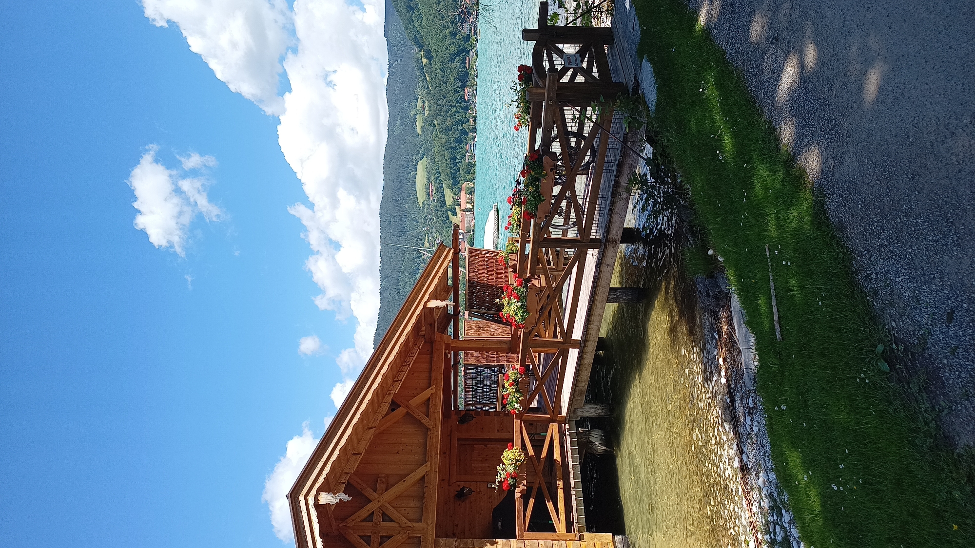 A boathouse on Lake Tegernsee, in Rottach-Egern, Germany, is captured from the perspective of someone standing on a gravel path. The boathouse, constructed of weathered wood, features a pitched roof and a covered porch extending over the water. A wooden walkway, adorned with planters full of red flowers, leads to the boathouse. The clear, turquoise water of the lake gently laps against the shore, where small stones are visible. Beyond the boathouse, the picturesque landscape of rolling green hills, dotted with buildings and trees, rises to meet a bright blue sky filled with scattered white clouds. The overall scene conveys a sense of serene natural beauty and rustic charm.