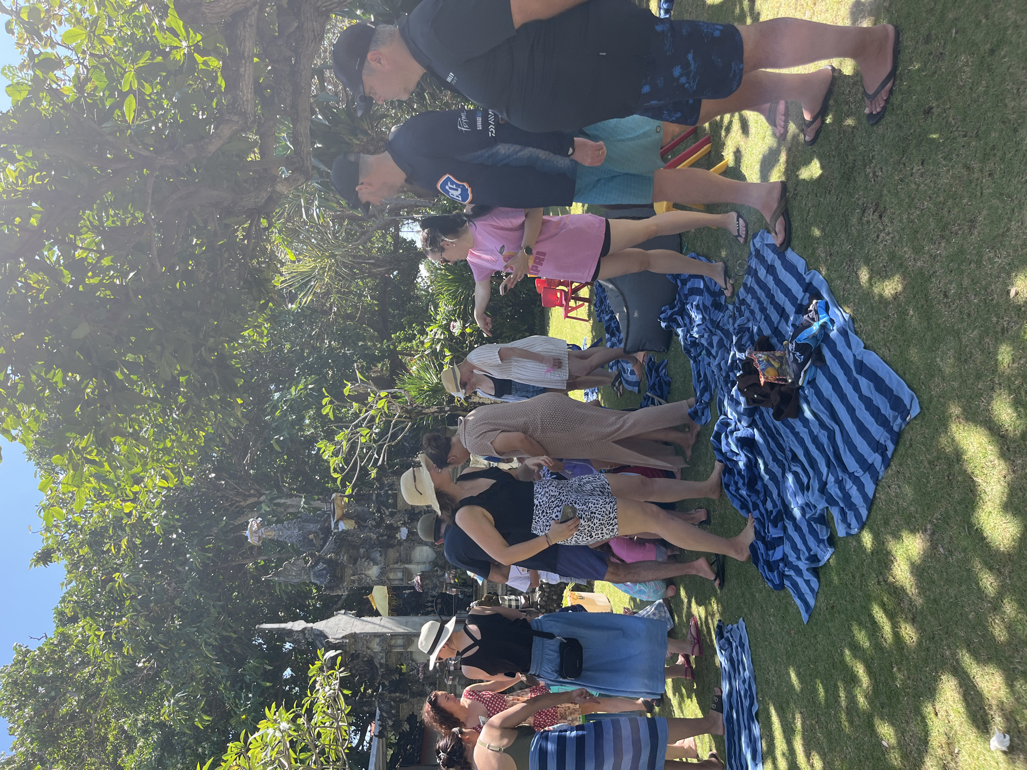 In Kuta Selatan, Indonesia, a group of approximately 15-20 people are gathered outdoors on a sunny day in a grassy area. The scene suggests a casual social gathering, possibly a beach or resort setting, given the presence of numerous striped blue beach towels laid out on the grass. Several individuals are wearing casual attire, including swimwear, shorts, and t-shirts, and some are wearing hats. The background features lush green trees and foliage, with a glimpse of what appears to be a decorative stone structure, possibly part of a temple or a garden feature. The overall atmosphere is relaxed and informal, with people standing, interacting, and some appearing to be looking at something or someone off-camera. The ground is covered in green grass, and the lighting indicates bright daylight.
