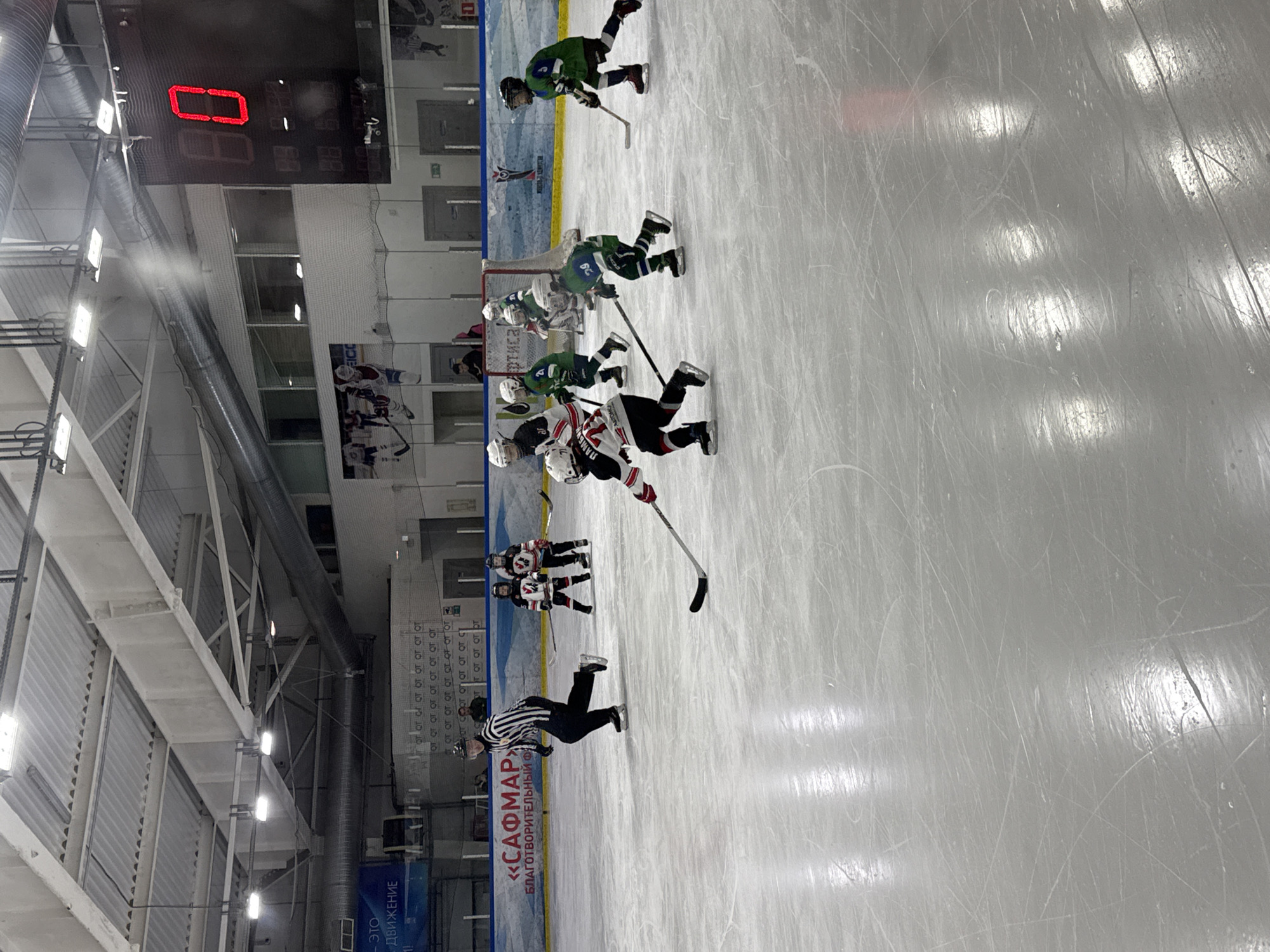 A hockey game is in progress at a rink in Izhevsk, Russia. The score on the digital scoreboard reads "0". Players in green and white uniforms are around the net. One player in red and white is challenging for the puck. A referee in black and white stripes is on the ice. There are two groups of players on the sidelines.
