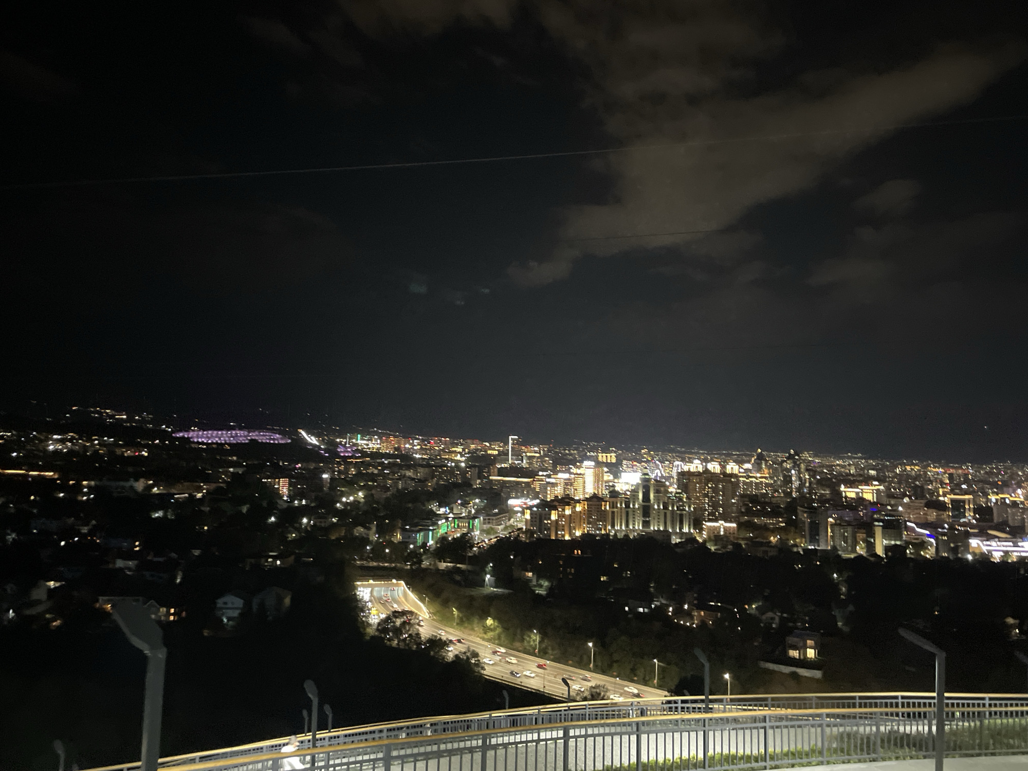 This is a night-time, elevated, wide-angle shot of Almaty, Kazakhstan. The foreground shows a metal railing with a light grey painted surface, supported by vertical posts and a yellow accent strip. Beyond the railing is a dark, sloped hillside.
In the mid-ground, a multi-lane highway is visible, with traffic lights and headlights from numerous vehicles. The road curves and disappears into a tunnel on the left side of the image. Trees and residential buildings with illuminated windows are scattered across the hillsides.
The background is dominated by the city skyline, illuminated by thousands of lights. Tall buildings with bright lighting are prominent in the center and right of the frame. The lights are a mix of white, yellow, and some purple, particularly in the distant left portion of the city. Clouds are present in the night sky, with some illumination from the city lights reflecting off them. A faint, thin horizontal line, possibly a wire or antenna, is visible across the sky. The overall impression is a sprawling, well-lit urban landscape under a dark, cloudy night sky.