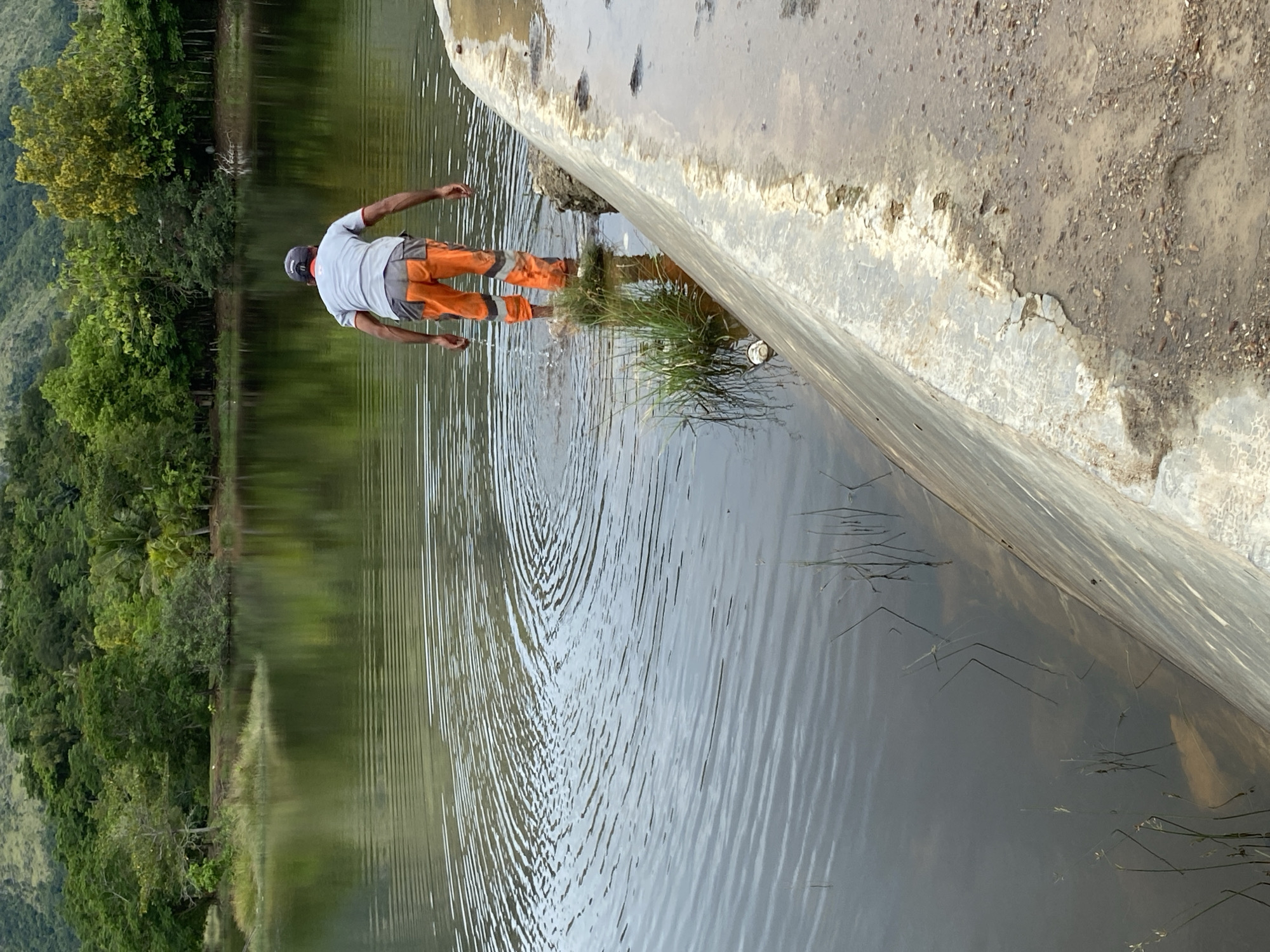 In Aneuk Paya, Indonesia, a person wearing a grey shirt, a dark baseball cap, and orange work pants with reflective stripes stands in shallow water near a concrete structure. The individual is facing away from the camera, with their hands lowered towards the water's surface, creating ripples. The environment consists of a body of water reflecting a lush green hillside covered in trees. The concrete structure appears to be a dam or retaining wall, with a rough, textured surface and some debris on the adjacent dry ground. The water is calm, with visible concentric ripples originating from the person's movement.