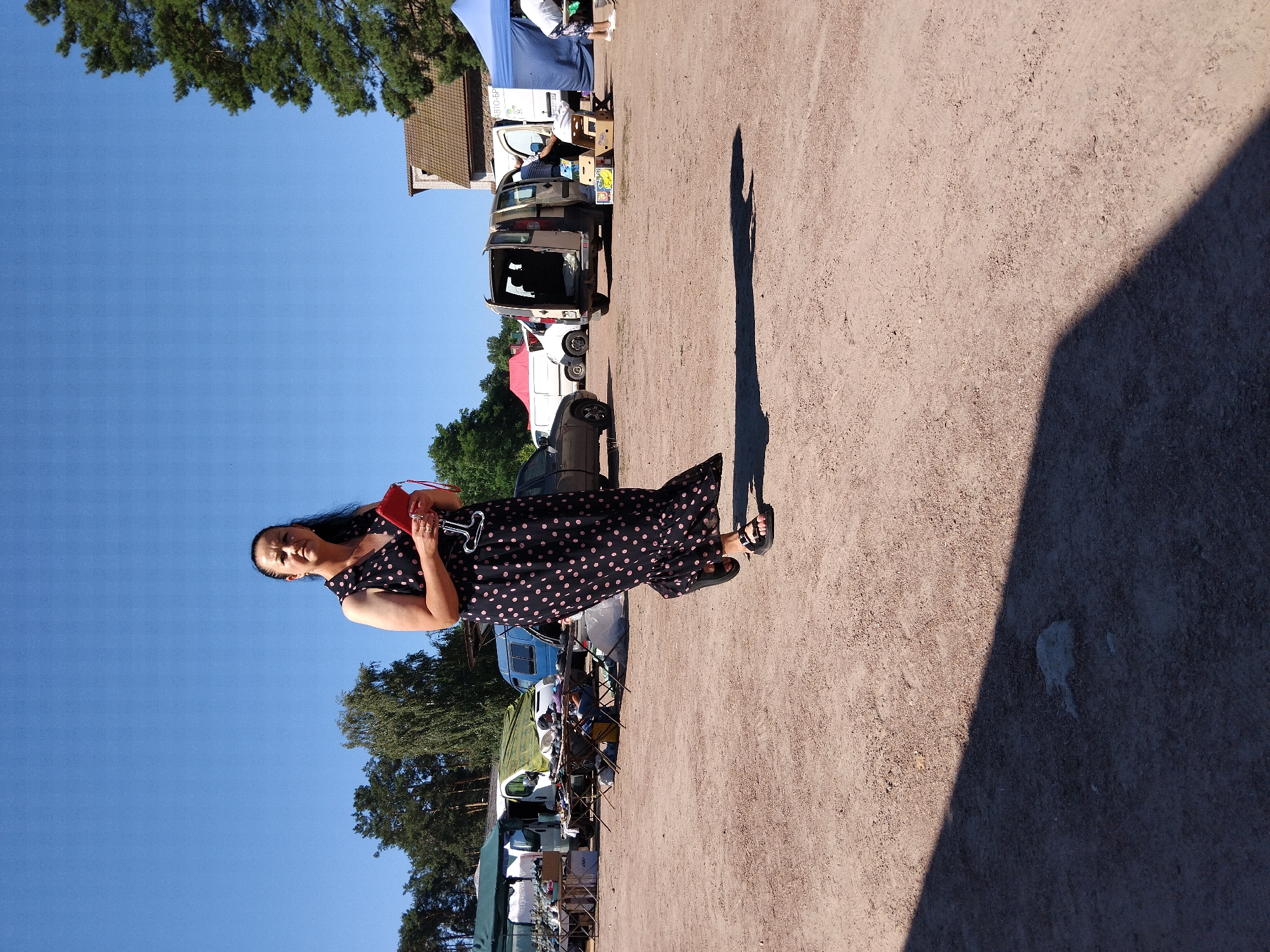 A woman in a black polka-dot dress is walking in an outdoor market on a sunny day. She is holding a red clutch and a glass. In the background, there are tents, vans, and trees. The ground is covered in dirt and gravel. The location is identified as Irshansk, Ukraine.