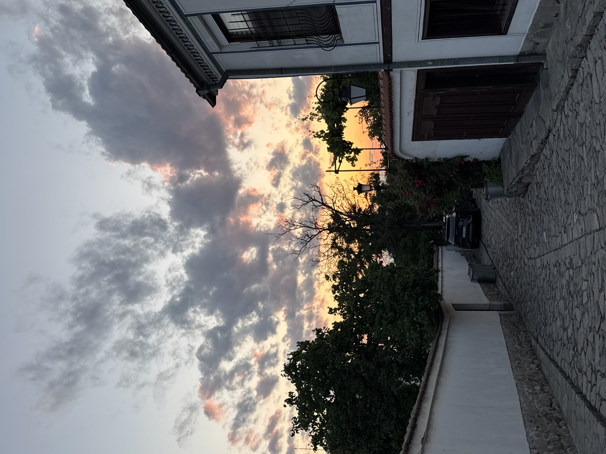 The image depicts a cobblestone street in the Old Town of Plovdiv, Bulgaria, captured during sunset. The sky, visible above the buildings and trees, features a gradient from pale grey-blue at the top to vibrant orange and pink hues near the horizon, interspersed with scattered dark grey clouds.
On the left side of the street, a long, whitewashed stone wall with a darker stone base runs parallel to the street. Above this wall, dense green foliage of trees is visible. Further along the wall, there is a dark, recessed entrance or doorway, followed by two metal trash bins. A dark-colored SUV is parked further down the street on the left.
The cobblestone street, made of irregular grey stones, slopes gently and curves towards the background.
On the right side of the street, a traditional Bulgarian Revival-style house stands prominently. It features white walls, dark window frames with decorative wrought iron grilles, and detailed blue patterns along its eaves and under the roofline. A dark brown double wooden door, possibly belonging to a garage or a separate entrance, is located on a lower section of the building or an adjacent structure. An ornate black street lamp with a white lantern is mounted on a pole, from which a leafy vine extends across a horizontal support. No individuals are present in the scene.