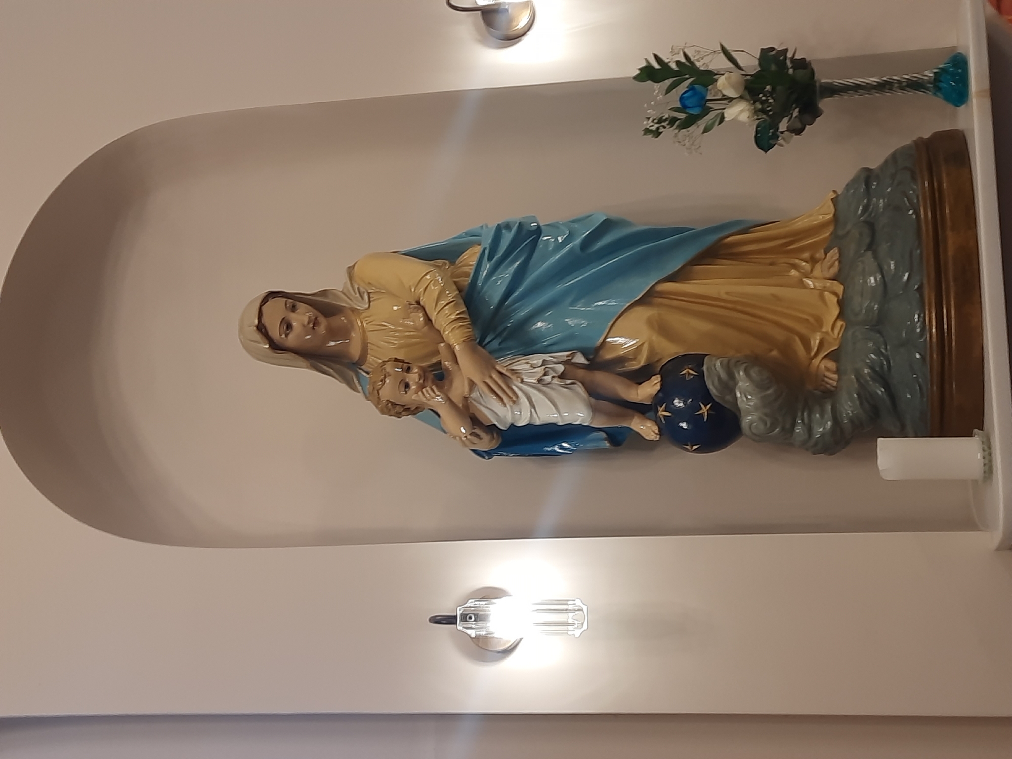 This media file captures a religious statue of the Virgin Mary holding baby Jesus, situated within an arched recess in an indoor setting. The statue depicts the Virgin Mary in a golden gown and a prominent blue mantle, gently cradling baby Jesus, who is dressed in white. The base of the statue features a gray, cloud-like formation, and a blue sphere adorned with golden stars.
Above and to the right of the statue, a vase containing artificial blue and white flowers adds a decorative touch. The scene is illuminated by two modern, bright light fixtures, one positioned above and another below the statue, casting a strong, direct light that creates reflections on the glossy surfaces. The walls of the recess and the surrounding area are a light, neutral color.
The image portrays a devotional scene, likely in a home or religious space in Kadıköy, Turkey. There is no discernible activity taking place, as it is a still depiction of statuary, and no visible text is present. The overall mood conveyed is serene and reverent.