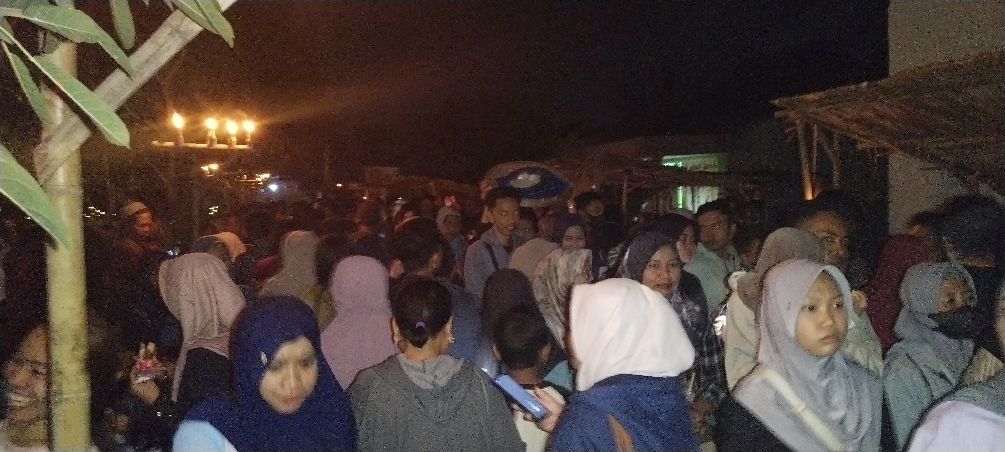 The image depicts a nighttime outdoor gathering of a crowd of people, many of whom are wearing headscarves. The setting appears to be a street or open area, with some structures visible in the background, possibly stalls or temporary shelters with thatched roofs. There are lights illuminating the scene, including what appears to be a candelabra with multiple lit candles in the upper left portion of the image. The people are closely packed together, suggesting a public event or festival. The quality of the image is low, with graininess and limited detail due to the nighttime conditions and resolution. The individuals are a mix of ages and genders, and their facial features are not always clearly discernible due to the lighting and distance. The overall atmosphere is that of a bustling community event. The location is specified as Keting, Indonesia.