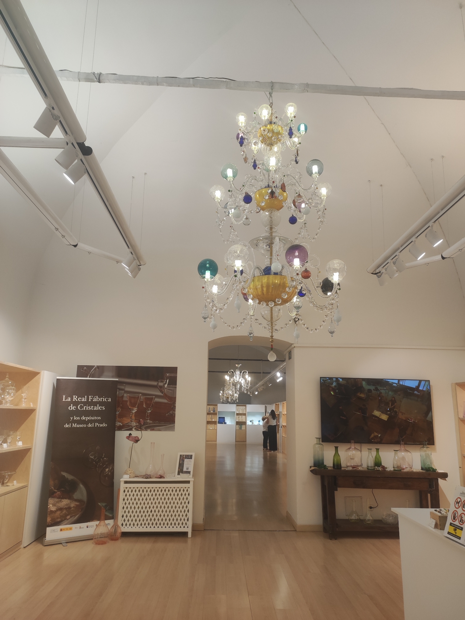 The image shows the interior of a museum or gallery in San Ildefonso, Spain, likely the La Real Fábrica de Cristales. The scene is dominated by a large, ornate crystal chandelier hanging from the ceiling. The room has white walls and a light-colored wooden floor.
On the left, there is a display of glass objects on shelves. A large banner with text "La Real Fábrica de Cristales y los depósitos del Museo del Prado" is visible, along with a white radiator cover. Two pink glass vases are on the floor in front of the banner.
In the background, a hallway leads to another part of the museum, with more displays of glass and a few people walking. To the right, a large television screen displays an image. Below the screen is a wooden table with several colored glass bottles. In the foreground on the right, part of a white counter or display is visible, with some signage. The overall impression is a well-lit exhibition space showcasing glass artistry.