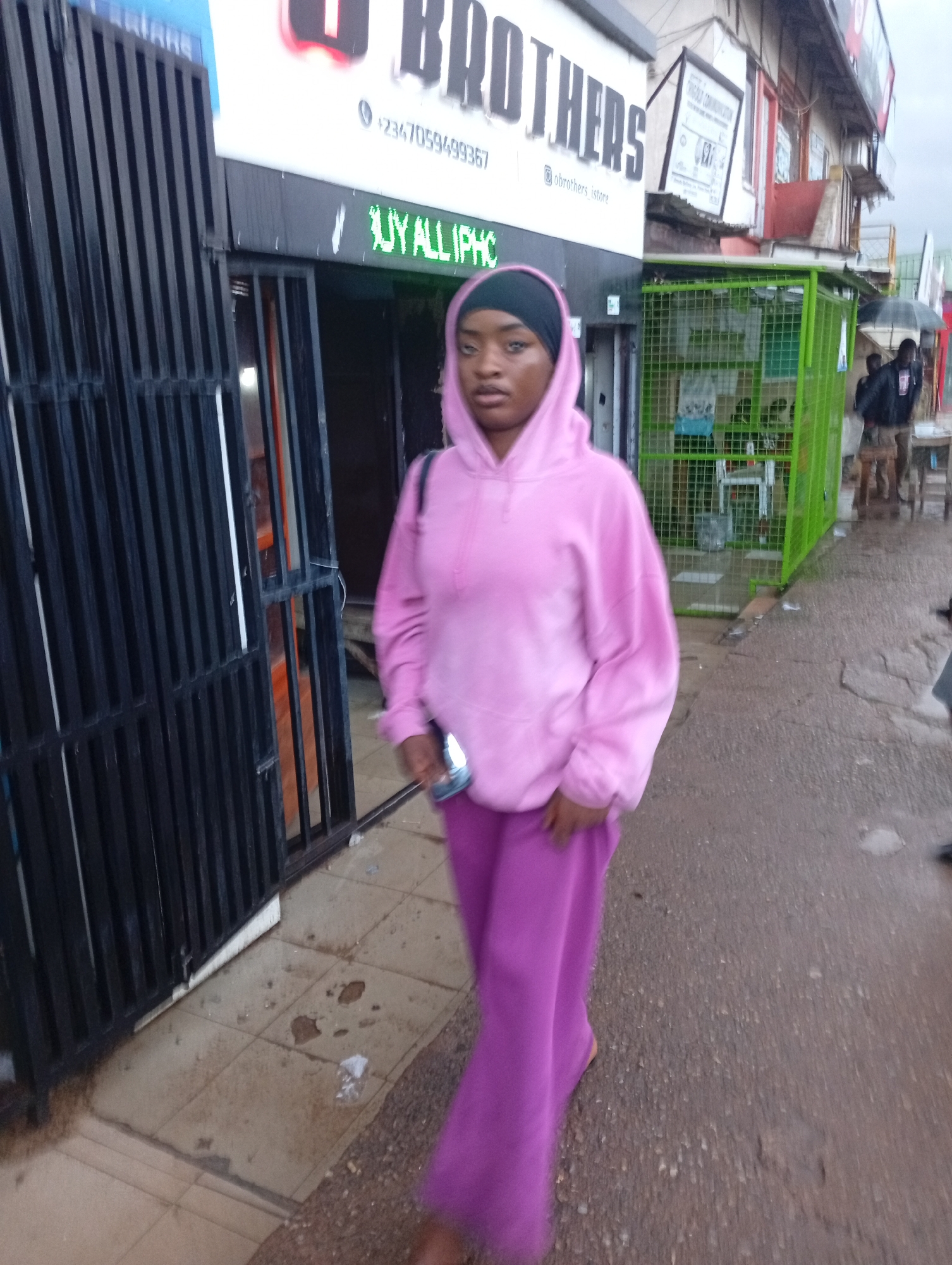 A young woman wearing a pink hoodie and purple pants walks on a wet paved street in Jos, Nigeria. She has a black head covering. The street is lined with shops, some of which have green metal cages in front of them. A sign above one shop reads "O BROTHERS" with a phone number and social media handle. Another sign in green LED lights reads "BUY ALL IPHC". The overall environment appears to be an urban street scene with multiple businesses and moderate pedestrian activity.
