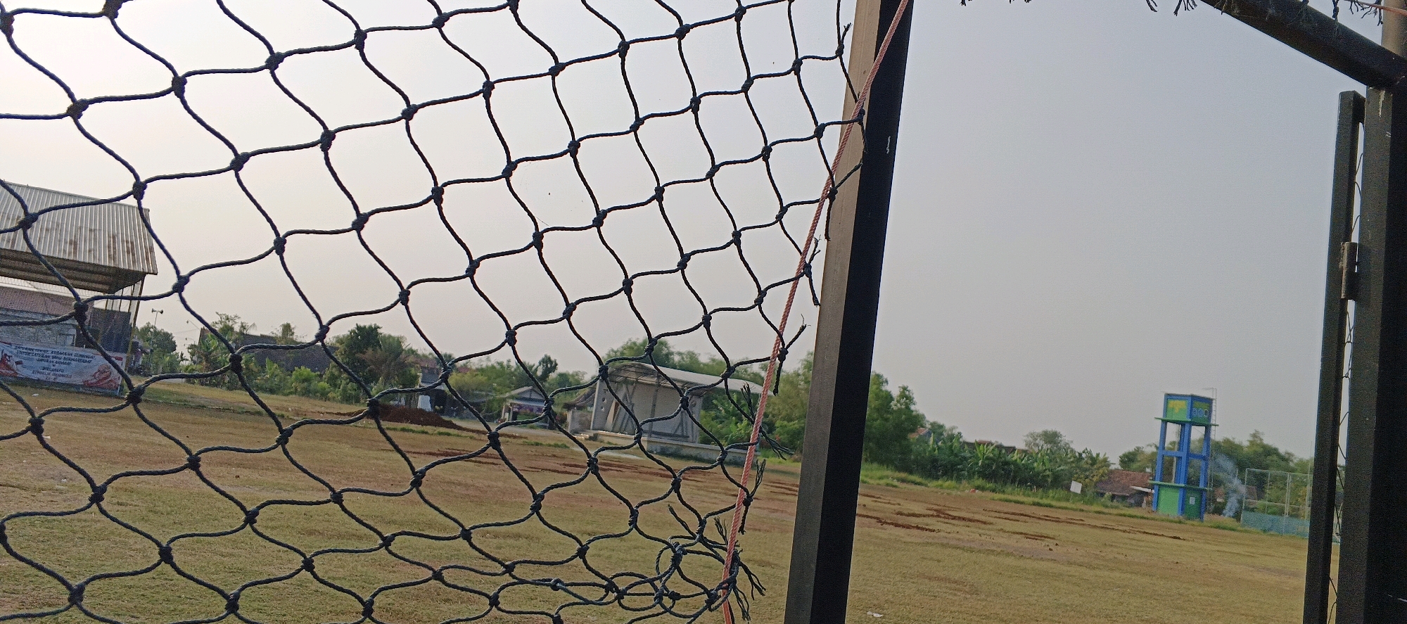 The image captures a view through a black mesh net, looking out onto an outdoor area. In the foreground, the net dominates the frame with its diamond pattern. Beyond the net, the scene unfolds. To the left, a large building with a corrugated metal roof stands on a slight incline. A banner with red and white elements is partially visible on the side of this building, suggesting a public or community space. Further into the background, a collection of houses and trees are scattered across the landscape, indicating a rural or semi-urban setting. On the right side of the image, a tall, blue and green water tower is visible against a pale, overcast sky. The ground in the mid-ground appears to be a field of dry, brown grass with patches of exposed soil. The overall impression is one of a typical Indonesian landscape on a hazy day.