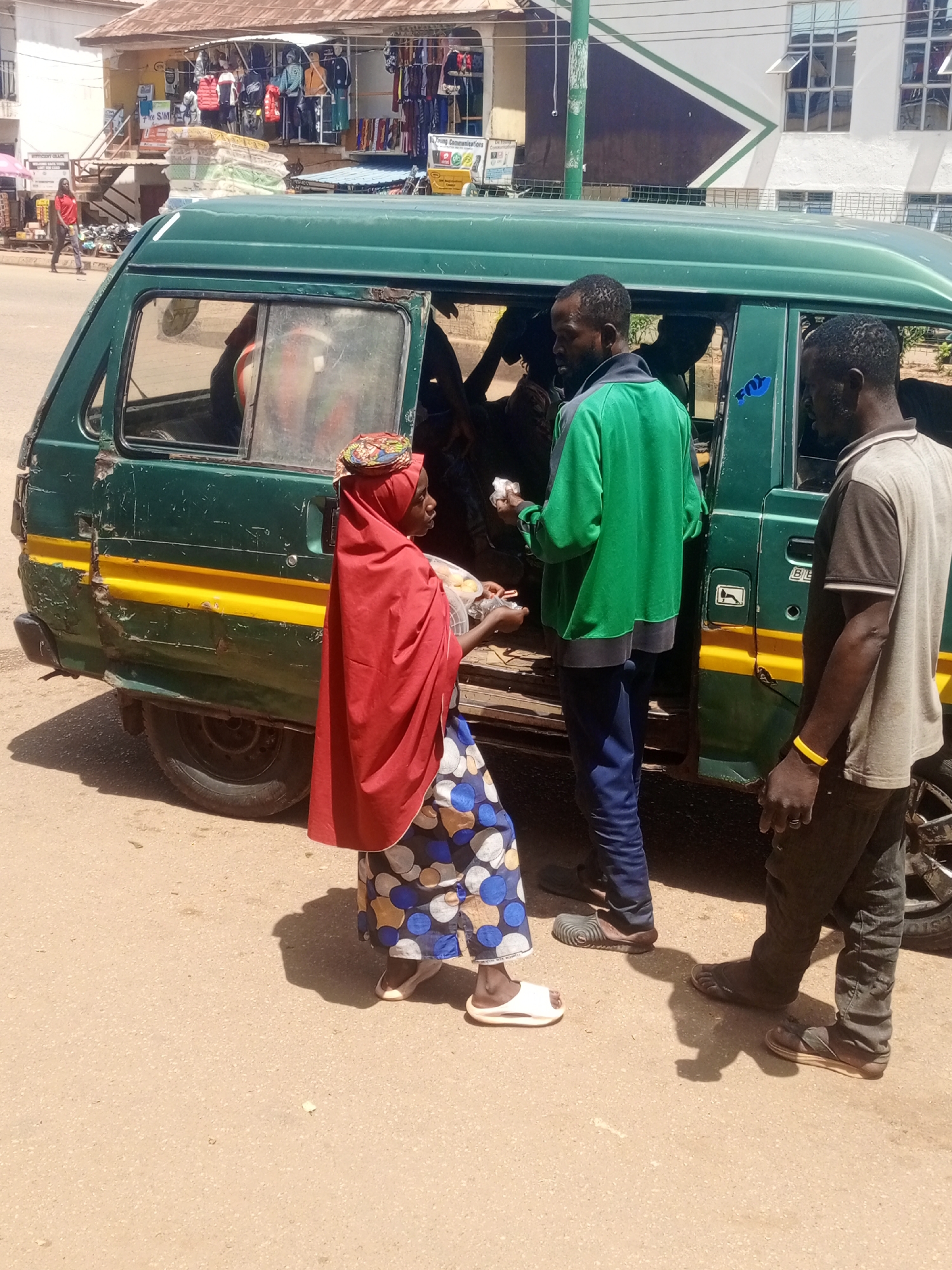 In Bukuru, Nigeria, under the bright afternoon sun, a group of people are engaged in an exchange by a parked green minibus. The minibus, with its weathered exterior and yellow stripe, appears to be a mode of public transportation. Three individuals are prominently visible. A woman in a vibrant red headscarf and a patterned blue and white skirt stands near the open passenger door, holding out items, possibly for sale. She wears white sandals. Facing her is a man in a bright green jacket and blue pants, reaching into the bus. Another man, wearing a grey t-shirt and dark pants with a yellow bracelet on his wrist, stands to the right, observing the transaction. His footwear consists of dark sandals.
Behind the minibus, shops with awnings display clothing and goods, suggesting a commercial area. The building in the background has multiple stories with large windows. The ground is paved with asphalt, and shadows indicate a clear, sunny day. The overall scene captures a moment of daily commerce and interaction in a bustling Nigerian town. There is no visible text clearly discernible in the image.