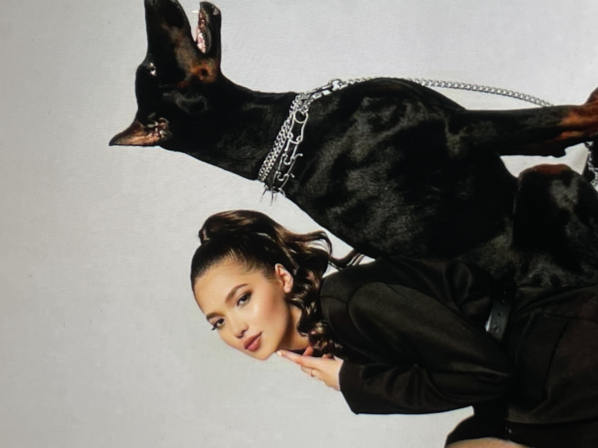 A woman with brown hair pulled into a high ponytail, wearing a dark outfit, is seen posing with a black Doberman dog wearing a spiked collar and chain leash. The background appears to be a plain, light-colored studio backdrop. The dog is positioned above and behind the woman, its head slightly raised. The location is Unknown, Unknown.