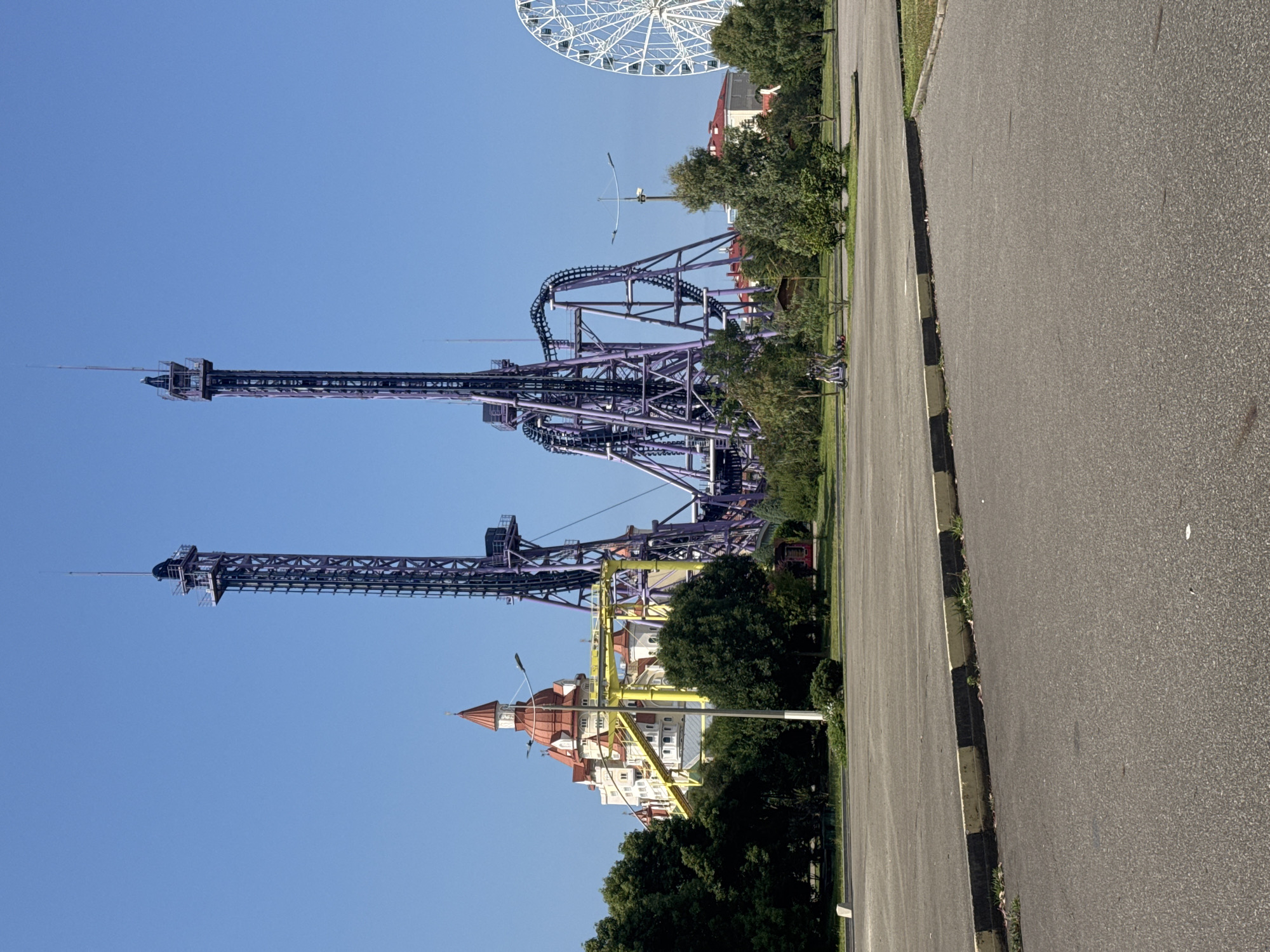 The image shows an amusement park with a clear blue sky overhead. In the center of the frame, two tall, purple roller coaster towers dominate the landscape, with the roller coaster tracks winding around them. To the left, a whimsical building with red, conical roofs resembles a castle. A yellow roller coaster track is visible in front of this building. On the far right, a white Ferris wheel is partially visible. The foreground consists of an empty asphalt parking lot with a yellow and black striped curb. Green trees and foliage are scattered throughout the park, providing a contrast to the man-made structures. In the distance, more buildings and trees are visible. A couple of electric scooters are parked in the middle ground on the grass. This amusement park is located in the city of Sirius, Russia.
