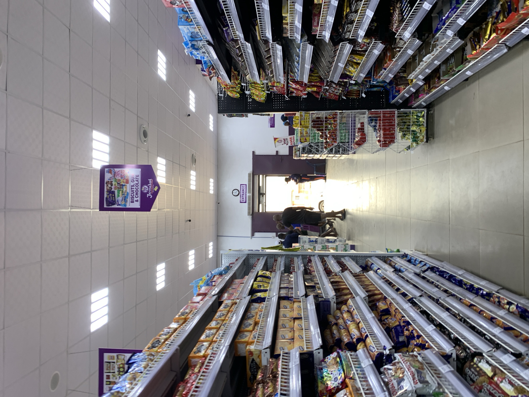 The photograph shows an interior view of a grocery store in Ikorodu, Nigeria. Fluorescent ceiling lights illuminate aisles lined with shelving units stocked with various packaged food products, mainly snacks and confectionery. A purple sign hanging from the ceiling advertises biscuits, gum, and chocolate. In the background, near the store's entrance, are several people: a man, a seated person, and two other individuals partially obscured by the brightness of the doorway. The store entrance is marked with an "ENTRANCE" sign. The floor is tiled.