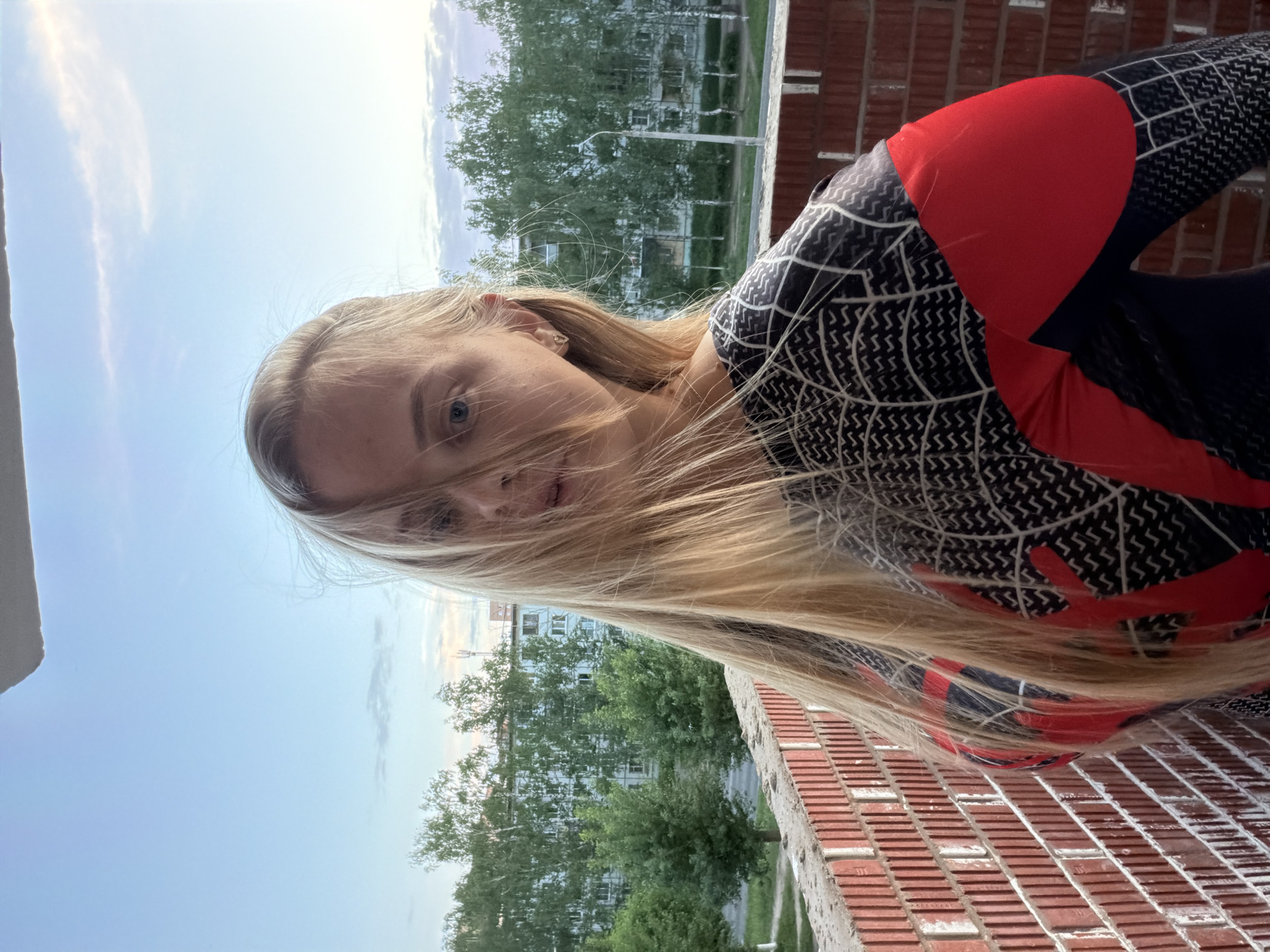 A young, fair-skinned woman with long blonde hair is on a balcony. She wears a form-fitting black and red Spider-Man-themed garment. The balcony has a red brick railing. Beyond the balcony, trees and a multi-story apartment building are visible, indicating an urban environment. It is outdoors and daytime. Based on the architecture, infrastructure, and clothing, the location is likely the city of Izhevsk, Russia.