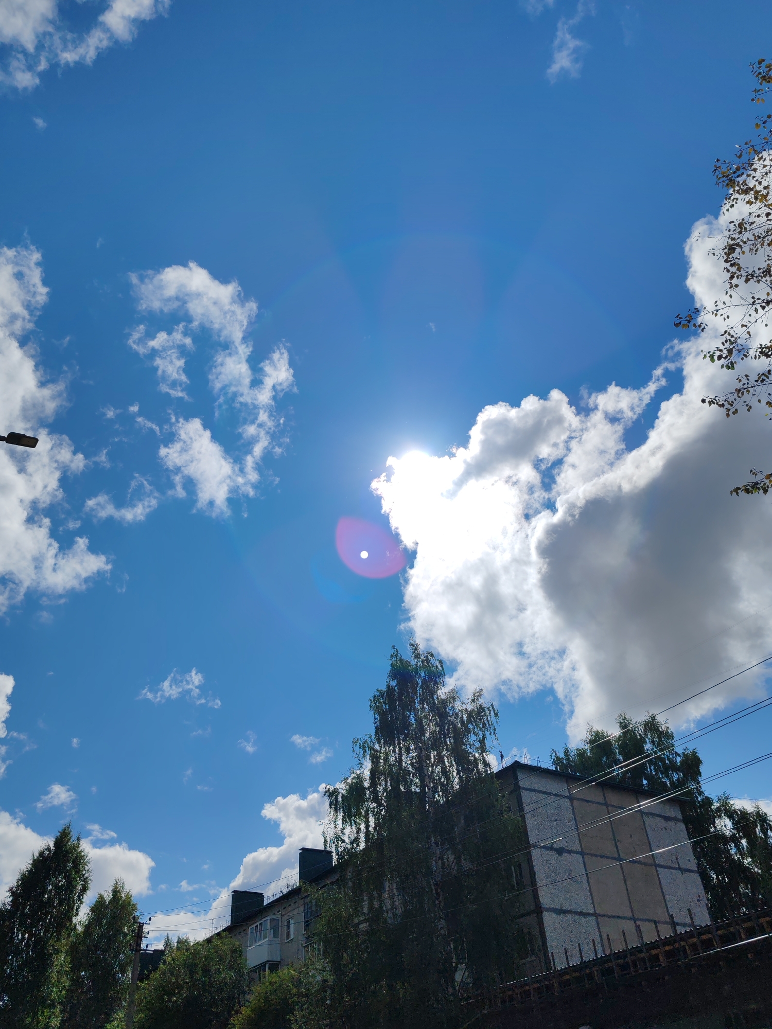The image captures a view of the sky and a building in Syktyvkar, Russia. The sky is a vibrant blue, dotted with fluffy white clouds. The sun is visible, creating a lens flare effect with a bright, circular halo. In the foreground, a multi-story apartment building is seen, with a light-colored facade and several windows. Trees with green foliage surround the building. Utility wires are strung across the scene. The overall impression is a bright, sunny day in an urban environment.
