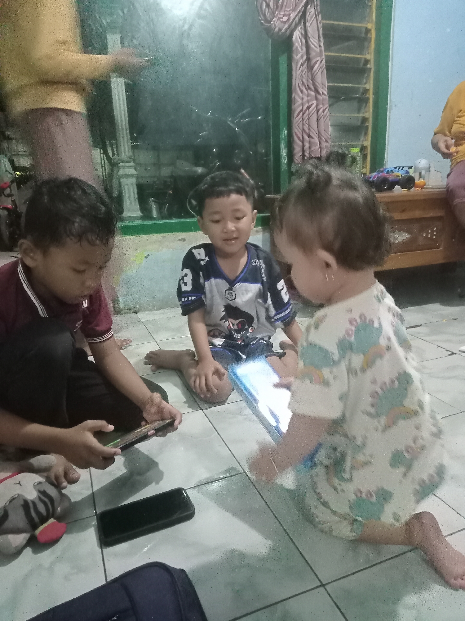The photograph captures a scene indoors, likely in a home located in Bungur, Indonesia. Three children are the central figures of the image. Two boys are seated on the floor, both engaged with electronic devices, presumably smartphones or tablets. The boy on the left is wearing a maroon collared shirt and dark pants, and he is focused on a phone in his hands. Beside him is a plush toy. The boy seated next to him is wearing a jersey with a number 3 and a graphic print. He is holding a tablet. In front of him, is a young child, possibly a toddler, wearing a light-colored outfit with a pattern of dinosaurs. They are also holding a tablet. The floor is tiled with light-colored square tiles. In the background, an adult is visible. The photograph suggests a casual, domestic setting.