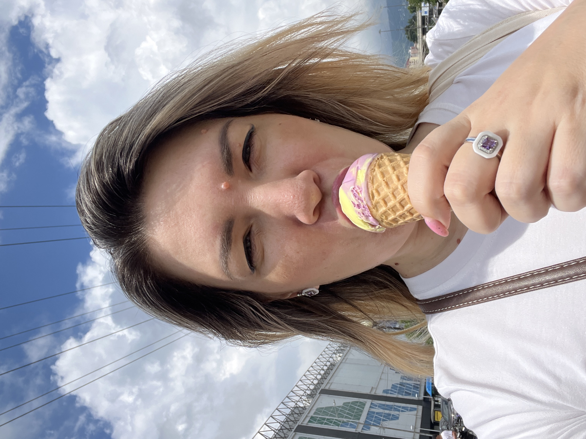 A woman is taking a selfie outdoors while eating an ice cream cone on a cloudy day. She is wearing a white t-shirt and a ring with a purple stone. The background shows a large building with a patterned facade, power lines, and some distant mountains. The location is identified as Sochi, Russia.