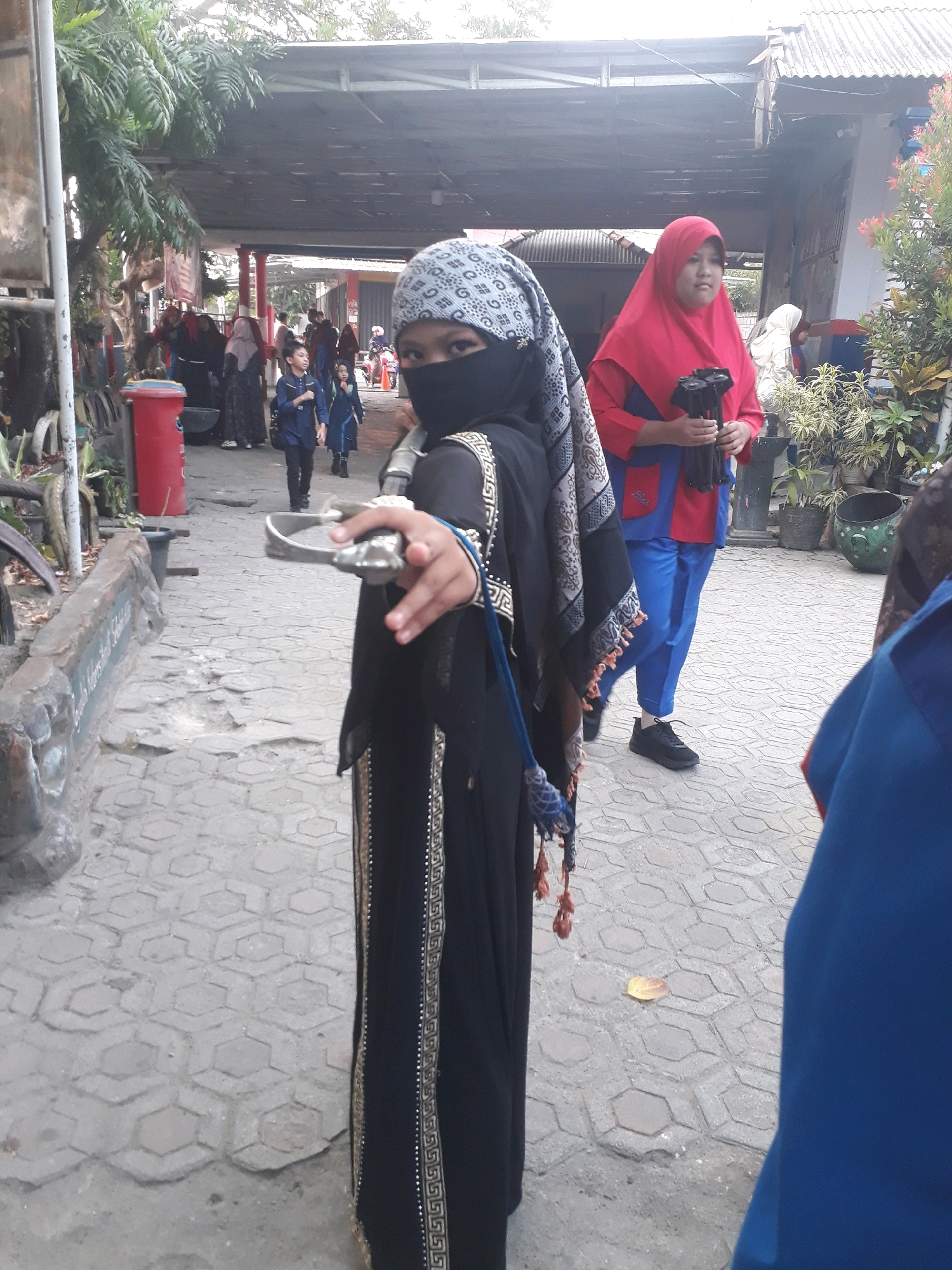 In Pangkalpinang, Indonesia, a person wearing a black abaya and a patterned headscarf stands in the foreground, holding a metallic object that resembles a sword hilt in their right hand. The individual's face is covered by a black niqab. Behind them, a woman in a red hijab and a blue and red garment holds a black object. Further in the background, several other people are visible, including children in blue outfits. The scene takes place outdoors in an area with a paved walkway and an overhead shelter structure. A red trash bin is present on the left side of the image. The environment appears to be a public space, possibly a school or community area, with some foliage and potted plants visible.