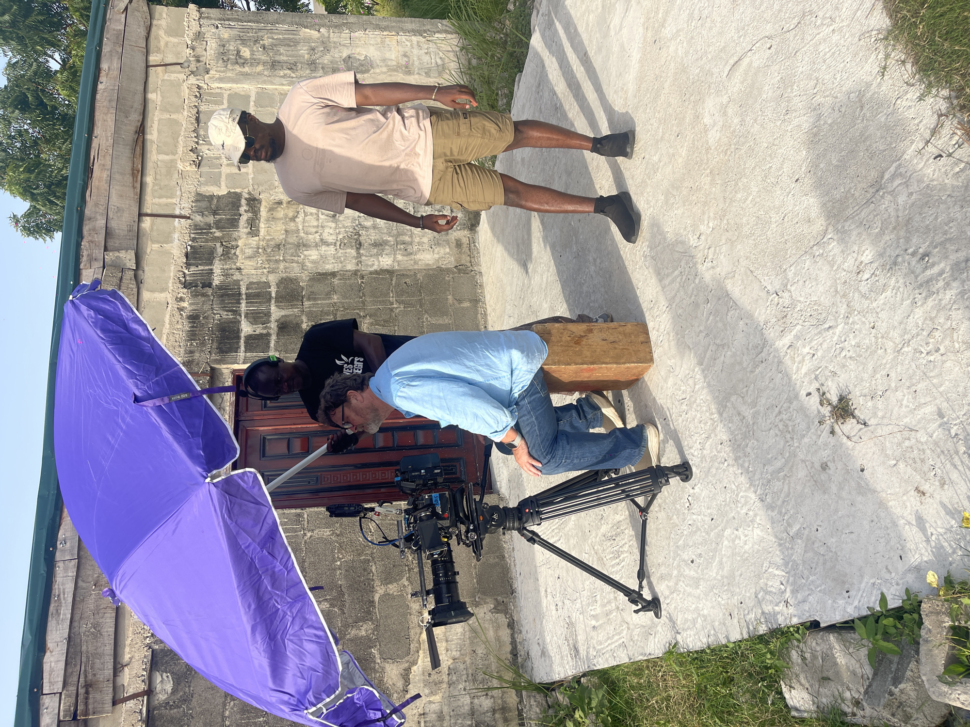 The image shows a film set with two men and a camera. One man is standing, wearing a tan shirt, khaki shorts, a white hat, and black boots. He is positioned on a concrete surface. The second man is kneeling behind a large film camera mounted on a tripod, with a wooden box between him and the camera. He is wearing a light blue shirt and jeans. Another person, possibly part of the crew, is partially visible behind the man kneeling, wearing a black shirt and headphones. A large purple umbrella or reflector is positioned to the left of the camera. The backdrop includes a building constructed from gray blocks. The location appears to be outdoors, with grass visible around the concrete surface.