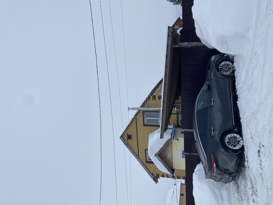 A forensic examination of the image reveals a winter residential scene in Мокино, Russia, under an overcast sky. A dark-colored sedan, consistent with a Kia Optima model, is parked amidst deep snow. The vehicle's exterior is visibly soiled with snow and road grime. Its front end is positioned near a large snowdrift, while the rear exhibits tire tracks in the snow leading away from it. The car is partially sheltered by a rustic wooden carport structure, which has snow accumulated on its roof.
In the background, a two-story house is visible. The lower section of the house is light yellow, with a small, snow-laden gable over what appears to be an entrance or bay window. The upper story is constructed from light brown or yellow brick, featuring multiple windows and a prominent silver-colored chimney rising from its snow-covered gabled roof. A dark brown corrugated metal fence separates the foreground from the main house structure. Utility lines are visible traversing the upper portion of the frame against the grey sky. Further in the background to the right, a portion of another red-brick house with a snow-covered roof is discernible. No individuals are present in the scene.