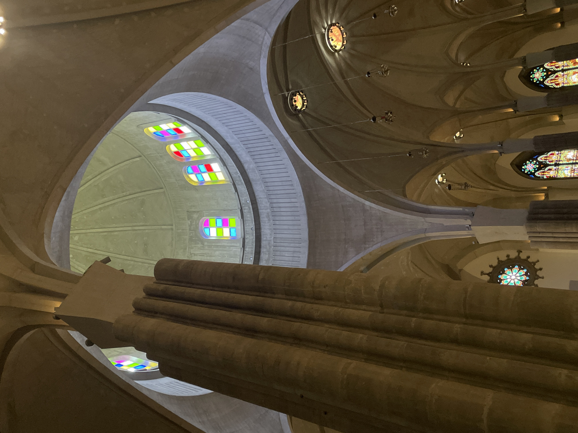 The image presents an upward-looking view inside a monumental architectural space, showcasing its intricate ceiling and upper structural elements. The scene is dominated by massive, smooth, and organically shaped columns and vaulted ceilings, likely constructed from concrete or light-colored stone. These structural elements branch out and converge, creating a complex, tree-like canopy effect overhead.
Several stained glass windows illuminate the interior. On the left side of the frame, a series of rectangular windows feature a modern, abstract design with bright, distinct blocks of primary and secondary colors (red, green, blue, yellow, magenta, cyan, white) arranged in a grid. Further into the distance and towards the right, other circular or rosette-shaped stained glass windows display more traditional, ornate patterns with a vibrant mix of colors.
Small, warm-toned light fixtures are subtly integrated into the ceiling vaults and along the structural arches, contributing to the overall subdued and majestic illumination. The architecture is characterized by sweeping curves, geometric precision, and a blend of modern and organic forms, creating a sense of grandeur and awe.
There are no people or specific activities depicted; the focus is entirely on the building's interior. The overall lighting is warm, suggesting an ambient or artificial light source complementing any natural light filtering through the stained glass. No visible text is present in the image. The location context provided is San Cristóbal De La Laguna, Spain.