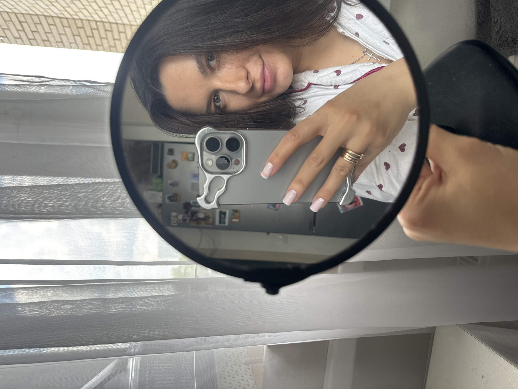 A person is taking a selfie in a round, black-rimmed mirror. The person has long dark hair and is wearing a white patterned shirt and a silver cross necklace. They are holding a grey smartphone with a triple-lens camera. The person is wearing several rings on their ring finger and has pink manicured nails. The background reflection shows a window with sheer white curtains and a glimpse of a refrigerator with magnets. The location is indicated as Akulovo (Gp Kubinka), Russia.