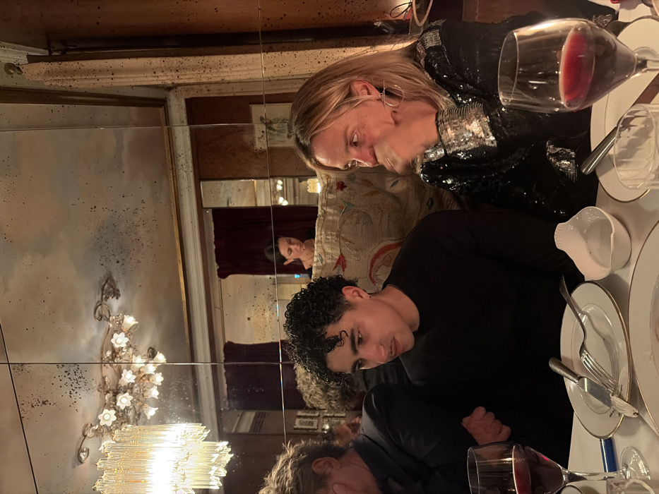 The image depicts an indoor scene within the Caviar Kaspia restaurant in Paris, France. Three individuals are seated at a dining table, observed in a dimly lit, opulent setting characterized by antique mirrors and chandeliers.
In the foreground, a blonde-haired woman with a large silver-toned hoop earring on her left ear is visible on the right, wearing a black top with silver sequin embellishments. Her right hand is positioned near her mouth, partially obscuring her face as she looks slightly to her left. To her immediate left, a man with dark, curly hair and a faint mustache wears a plain black long-sleeved shirt. He has a small earring in his left ear and is looking downwards and slightly to his left. A third individual, a man with reddish-brown hair and a dark jacket, is partially visible on the far left.
The table is covered with a white tablecloth and features two wine glasses containing red wine. In front of the curly-haired man, a stack of white plates with gold rims holds a knife and a fork. A small, white, shell-shaped ceramic dish rests between the two foreground individuals.
The background consists largely of large, segmented antique mirrors exhibiting significant foxing and discoloration, reflecting elements of the room. These reflections include an ornate, classical chandelier with multiple floral-shaped lights and a dark-haired woman in a dark top, looking downwards, seated or standing further back in the room. A modern chandelier, constructed from vertical glass rods, is visible on the upper far left, emitting a warm glow. Plush banquette seating with an intricate, faded embroidered pattern is visible along the wall, alongside dark wood paneling and a deep red curtain, contributing to the high-end Parisian restaurant ambiance.