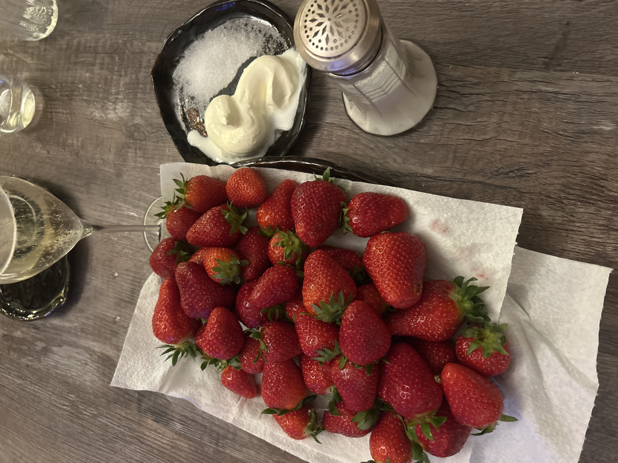 On a dark gray wood-grain surface, an overhead shot depicts an assortment of foods and beverages. A rectangular paper-towel-lined tray holds numerous red strawberries with green stems. To the left of the strawberries is a small container of white sparkling wine, and in the upper left corner are two small clear glasses, one empty and one slightly filled. A small black plate contains white granulated sugar and a dollop of white cream. Next to the plate is a silver salt shaker. The location is Unknown, Unknown. No people are visible. The image suggests a snack or dessert arrangement.