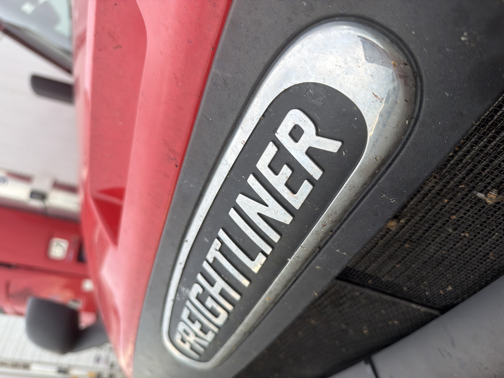 The image is a close-up, low-angle, outdoor photograph of the front grille and hood emblem of a red Freightliner semi-truck. The emblem, which reads "FREIGHTLINER" in silver raised letters within a chrome-finished oval, is the central focus. The truck appears to be parked, possibly in a lot or on a street in Murfreesboro, United States of America. The foreground shows a black mesh grille, partially obscured by the hood. The red paint of the hood is visible, showing some dust and minor scuffs. The background is slightly blurred, showing more of the truck's cab and possibly other vehicles or structures. The lighting suggests it is daytime. There are no people visible in the image.