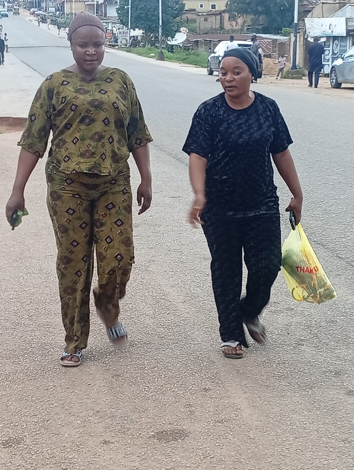 Two women are walking on a paved road, likely in Jos, Nigeria. The woman on the left wears a brown headscarf, a patterned green and brown top, and matching patterned trousers. She is holding a small green object in her left hand and wearing sandals. The woman on the right wears a black headscarf, a black textured top, and black trousers. She is carrying a yellow plastic bag in her right hand. The road appears to be a city street with buildings and vehicles visible in the background. The sky is overcast.