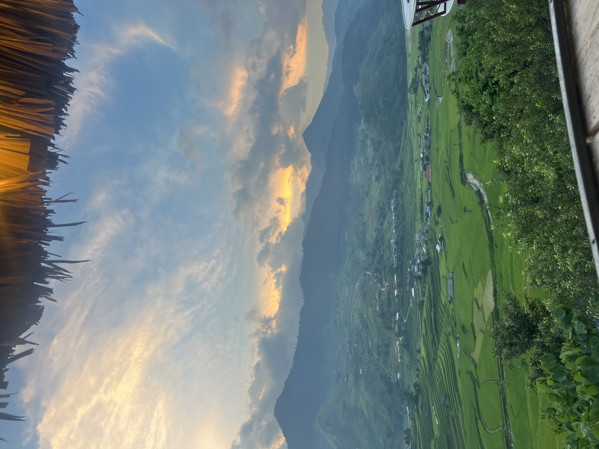 The image captures a picturesque landscape in Tu Le, Vietnam, likely during sunrise or sunset. In the foreground, a thatched roof structure is visible at the top, partially obscuring a vibrant sky filled with wispy clouds tinged with orange and yellow hues. Below the sky, a breathtaking vista of terraced rice paddies unfolds, presenting a rich tapestry of green hues. The paddies are meticulously sculpted into the rolling hills, showcasing the agricultural ingenuity of the region.
Nestled within the verdant landscape are small villages, characterized by clusters of houses with red roofs, blending harmoniously with their surroundings. A winding river or stream meanders through the rice fields, adding a touch of tranquility to the scene. The mountains in the background rise majestically, their peaks softened by a gentle haze. The overall impression is one of serene natural beauty and rural charm, characteristic of the mountainous regions of Vietnam. No people or animals are visible in the frame.