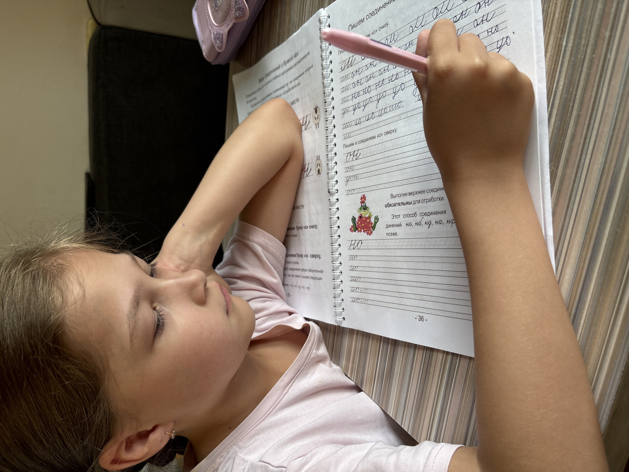 The image depicts a young girl, appearing to be of Eastern European descent, engaged in writing in a notebook. She is positioned at a table, with her left arm resting on it and her head tilted downwards as she concentrates on her work. She is wearing a light pink t-shirt. In the foreground, a pink pen is held by her right hand, poised over the notebook. The notebook contains handwritten text and line guides, suggesting it is an educational workbook, possibly for practicing calligraphy or penmanship. The environment appears to be a domestic setting, with a dark, upholstered object visible in the background, possibly a sofa or chair. A light purple pencil case, adorned with a cat's face design, is also present on the table. The lighting suggests an indoor setting, potentially during daylight hours. The girl's facial expression indicates focus and perhaps a slight furrowing of her brow. The table surface has a wood-grain pattern. There is no visible indication of the specific location being Bratsk, Russia.