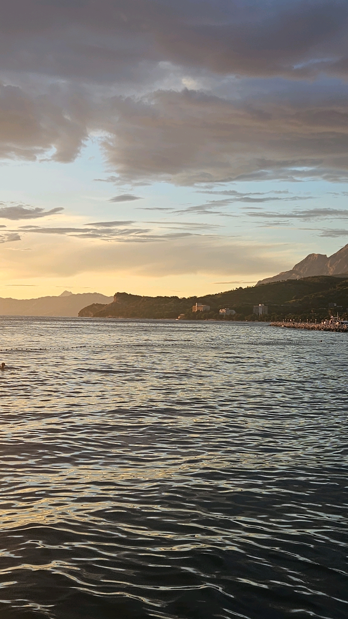 This is a vertical, eye-level shot of the sea and coastline in Tučepi, Croatia at sunset. The sky is a mixture of dramatic clouds in shades of gray, purple, and pink, with patches of pale blue and golden light. The sun is low on the horizon, casting a warm glow on the water and the distant hills.
The sea is calm, with small, rippling waves that reflect the colors of the sky. In the foreground, the water is a deep, dark blue, with streaks of golden light on the surface. A person can be seen swimming on the left side of the frame, their head and shoulders breaking the surface of the water.
In the middle ground, the coastline is lined with trees and buildings, including what appear to be hotels. The hills behind the coastline are covered in greenery. The horizon is marked by the silhouettes of mountains. A line of buoys or markers stretches across the water. The scene conveys a sense of tranquility and natural beauty.