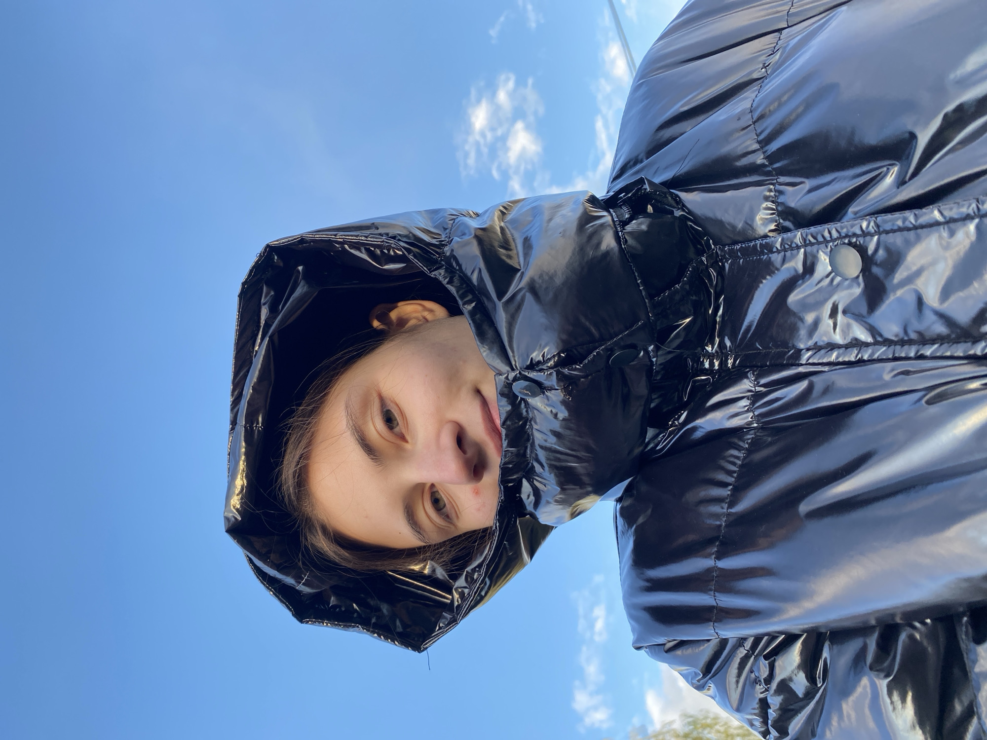 A young woman, viewed from below, is wearing a black, shiny puffer jacket with a hood. Her face is visible within the hood. She has brown hair, brown eyes, and fair skin. The background is a blue sky with scattered white clouds. The location is indeterminable from the image, and therefore not possible to discern its existence in the city of Votkinsk, Russia.