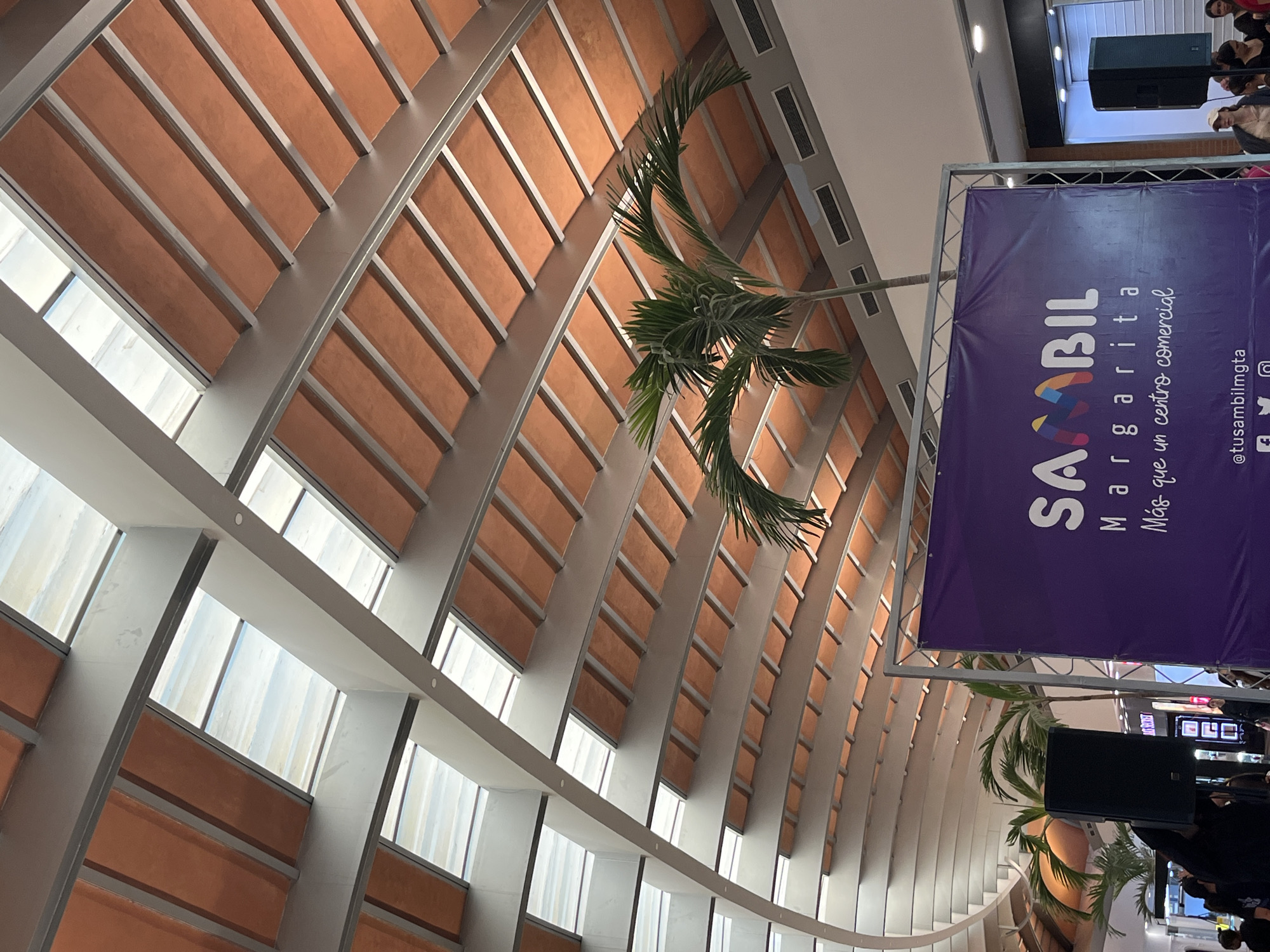 This media file captures the interior of a modern building, specifically identified as Sambil Margarita, a shopping mall in Pampatar, Venezuela, as indicated by a prominent purple banner. The scene is dominated by a distinctive architectural ceiling featuring a series of curved or arched light-colored beams alternating with reddish-brown panels. Integrated into this structure are numerous elongated rectangular windows that allow natural light to filter into the space, suggesting it is daytime.
Several palm trees are visible, with their fronds extending downwards from above, enhancing the tropical or outdoor-like ambiance within the indoor setting. On the right side of the image, a large purple banner hangs vertically. Text on the banner reads "SAABIL Margarita" (likely "SAMBIL"), followed by the slogan "Más que un centro comercial" (More than a shopping mall). Below this, social media handles including "@tusambilmgta" and icons for Facebook, Twitter, and Instagram are present.
Near the banner and further into the background, a large black speaker or sound equipment is visible. Scattered groups of people can be seen in the upper and lower right sections of the image, some gathered near what appears to be a stage or event area, suggesting an ongoing public event or a bustling environment within the mall. The overall impression is one of a spacious, well-lit, and architecturally interesting commercial center designed to incorporate natural elements.
