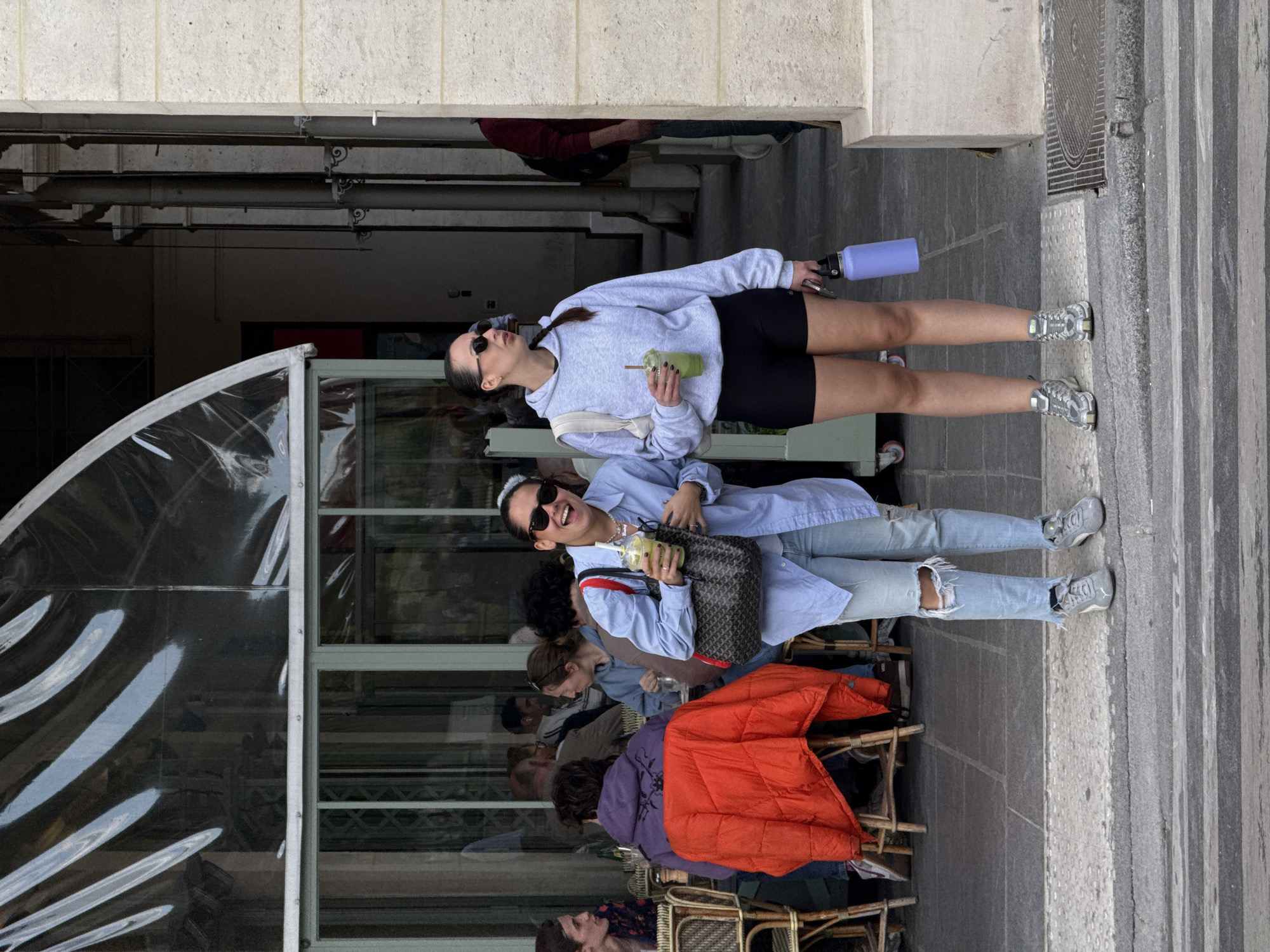 A full-shot, daylight photograph shows two young women standing on a paved sidewalk next to an outdoor cafe seating area in an urban setting, likely Paris, France. The image is rotated 90 degrees clockwise.
In the foreground, the woman on the left wears a light blue button-down shirt, ripped light-wash jeans, and grey sneakers. She has sunglasses on her head, a black shoulder bag with a distinct pattern (possibly Goyard), and is holding a clear cup with a green drink and a straw. She is smiling broadly and looking towards the camera.
The woman on the right wears a light grey hooded sweatshirt, black athletic shorts, and grey sneakers. She has sunglasses, a white cross-body bag, and is holding a similar clear cup with a green drink. She also holds a purple water bottle. Her expression is more neutral as she looks away to the right.
In the background, several people are seated at small wicker-style cafe tables, mostly obscured by glass and metal enclosures. One person is visible wearing a bright orange jacket draped over a chair, and another is in a purple hoodie. The ground is made of grey stone pavement, with a grated manhole cover and textured pedestrian strips visible on the right. Above the street level, a concrete structure and dark building elements are visible. The scene appears to be an everyday moment, possibly a break or leisure time. No discernible text is visible in the image.