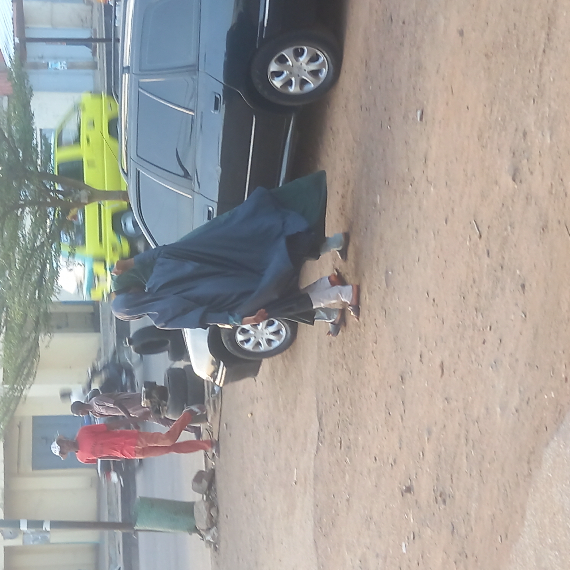 The image depicts a street scene in Dadi Riba, Nigeria. A black car is parked on a dirt-covered area. A person wearing a blue robe is walking next to the car. In the background, there is a yellow vehicle and a building. Two other people are walking in the distance. A motorcycle and other vehicles are visible. The environment appears to be urban or semi-urban.