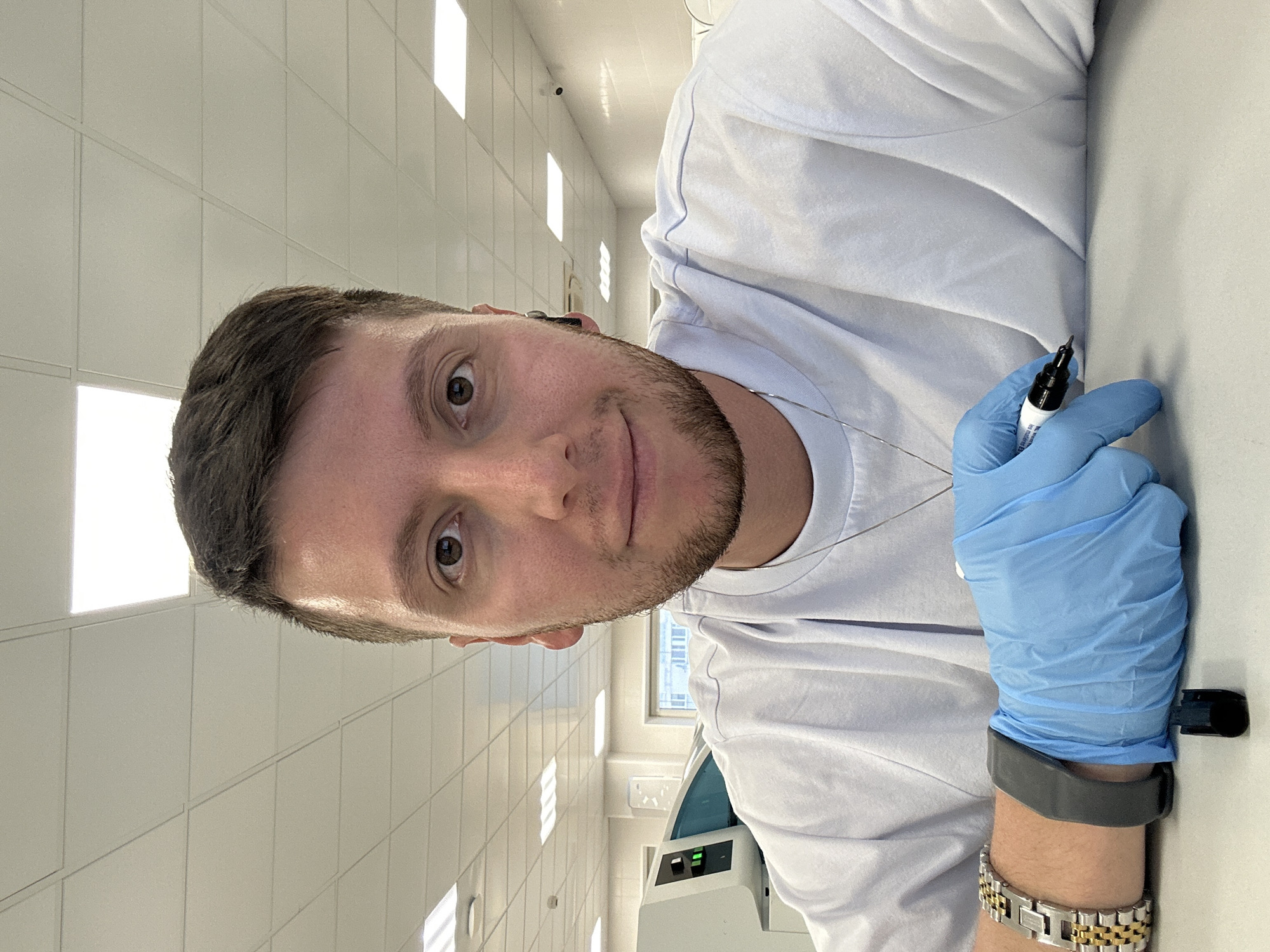 A man with short brown hair and a trimmed beard is holding a black marker in his gloved right hand. He wears a light gray smartwatch and a silver and gold bracelet on his right wrist. He is wearing a white t-shirt. The background shows a room with white tiled walls and ceiling, illuminated by rectangular ceiling lights. A piece of laboratory equipment with a green light is visible. The location is likely indoors but the city of Moscow, Russia cannot be determined from the visual information.