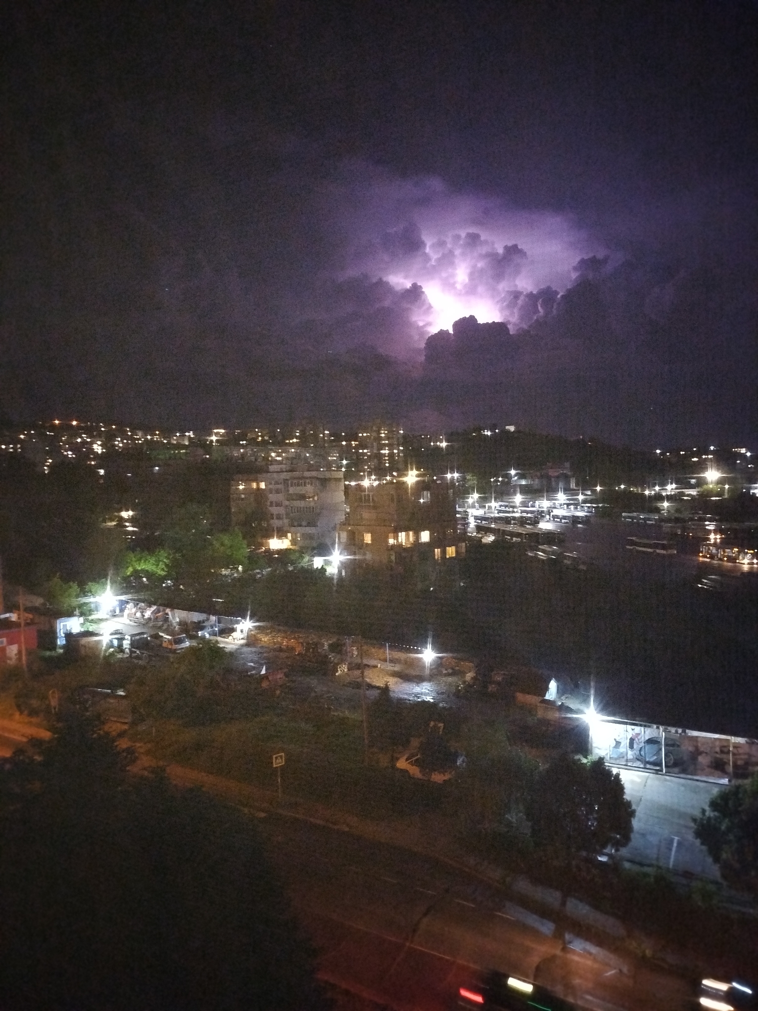The image shows a nighttime cityscape in Varna, Bulgaria, dominated by a thunderstorm in the upper right. Lightning illuminates dark storm clouds. Below, a densely populated urban area stretches across the frame. The scene features streetlights, building lights, and vehicle headlights, including blurred trails from moving cars on a road in the lower portion of the image. Several buildings and trees are visible in the foreground. A parking area or a construction site with parked vehicles and equipment is situated in the mid-ground. The overall environment is dark and rainy.