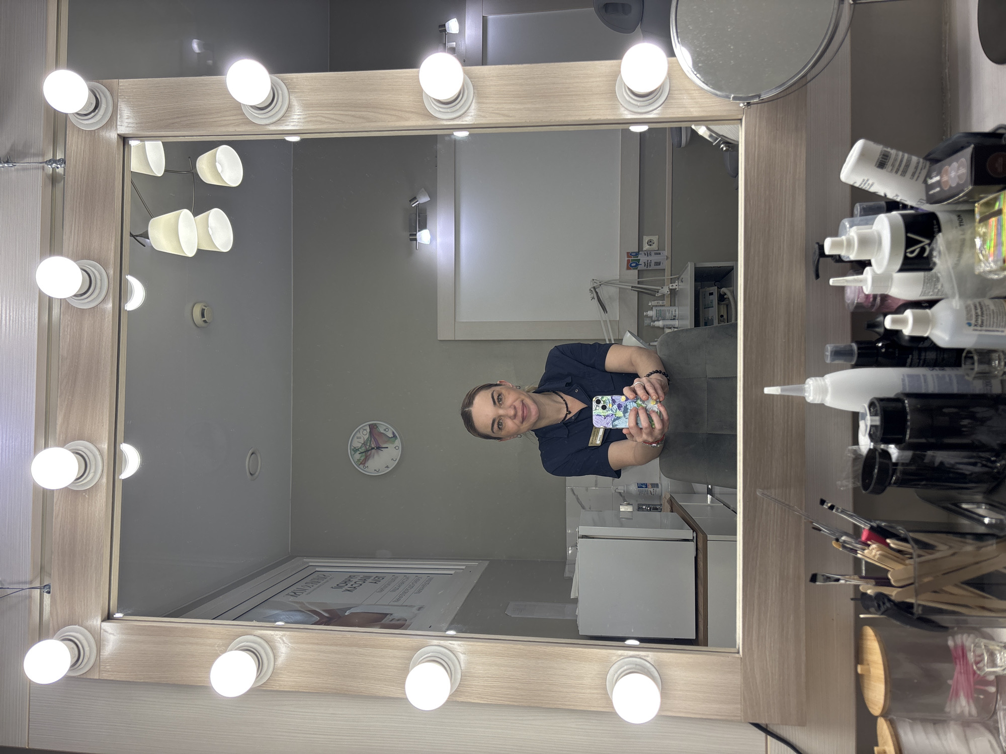 An interior shot depicts a woman taking a selfie in a large, brightly lit vanity mirror, likely within a beauty salon or aesthetic treatment room. The mirror has a light-colored wooden frame with multiple spherical light bulbs illuminating its perimeter. The woman, reflected in the mirror, is smiling and holds a smartphone with a colorful, patterned case. She wears a dark collared shirt, possibly a uniform, with a name tag or badge attached.
To the right of the mirror, a shelf is densely packed with various beauty products in bottles, brushes, wooden applicators, and cotton buds, suggesting professional use. The room reflected in the mirror behind the woman includes a wall clock with a colorful face displaying approximately 10:45, ceiling lights, a small fire alarm, a white cabinet, and a grey upholstered treatment chair. A smaller, circular magnifying mirror is also attached to the main mirror frame.
Visible text includes "SV" on one of the product bottles and partial text "MY LUXURY" along with Cyrillic script on a poster or sign reflected on the wall, possibly an advertisement. The overall atmosphere suggests a professional and well-equipped beauty-related establishment.